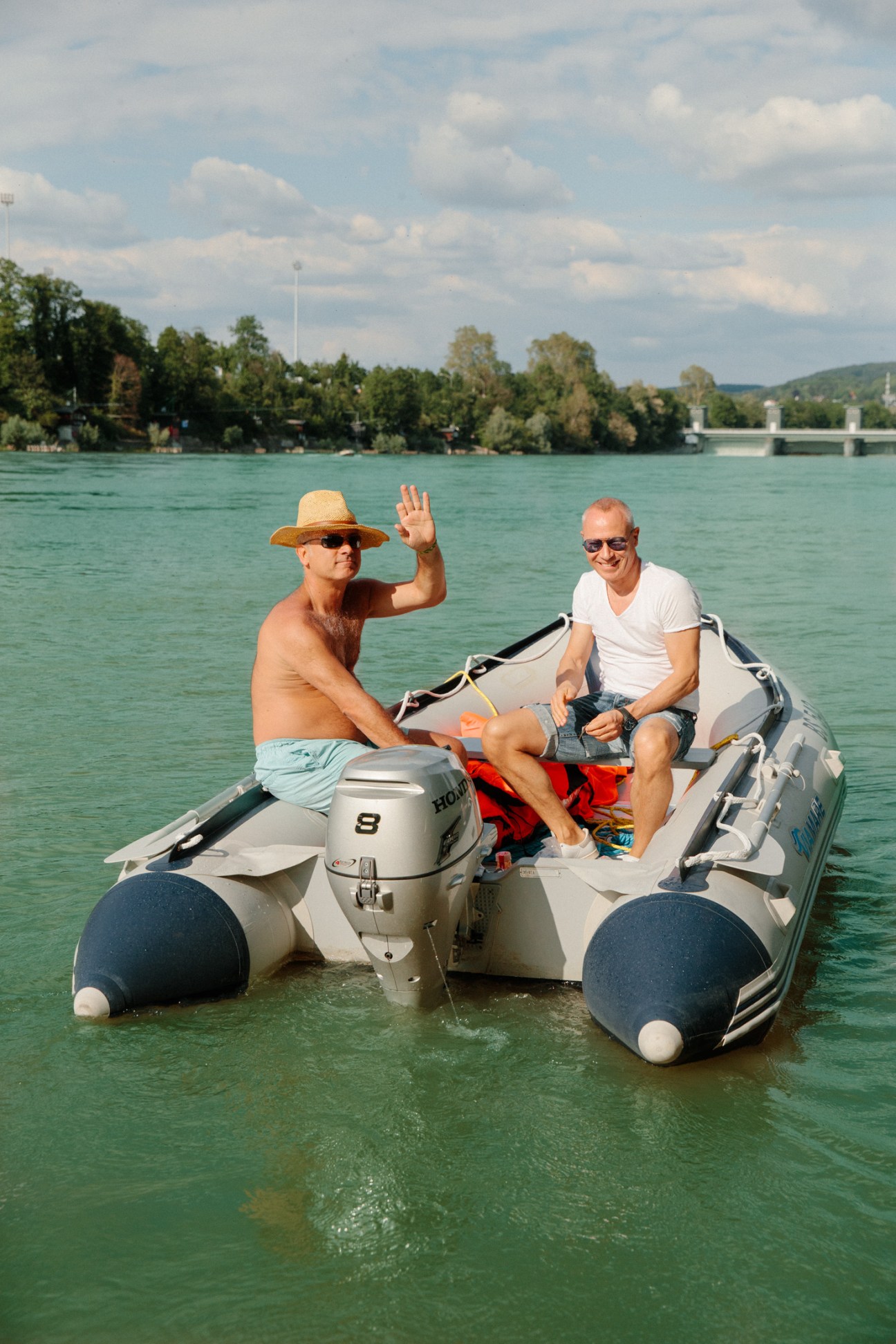 Two men in a small motorboat on turquoise Rhine river water with forested banks and bridge visible behind.