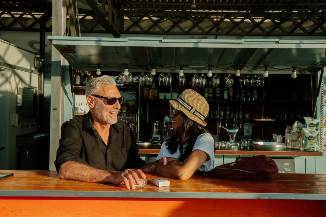 Two people chatting at the bar counter at Rheinbad Breite with hanging glassware visible behind.