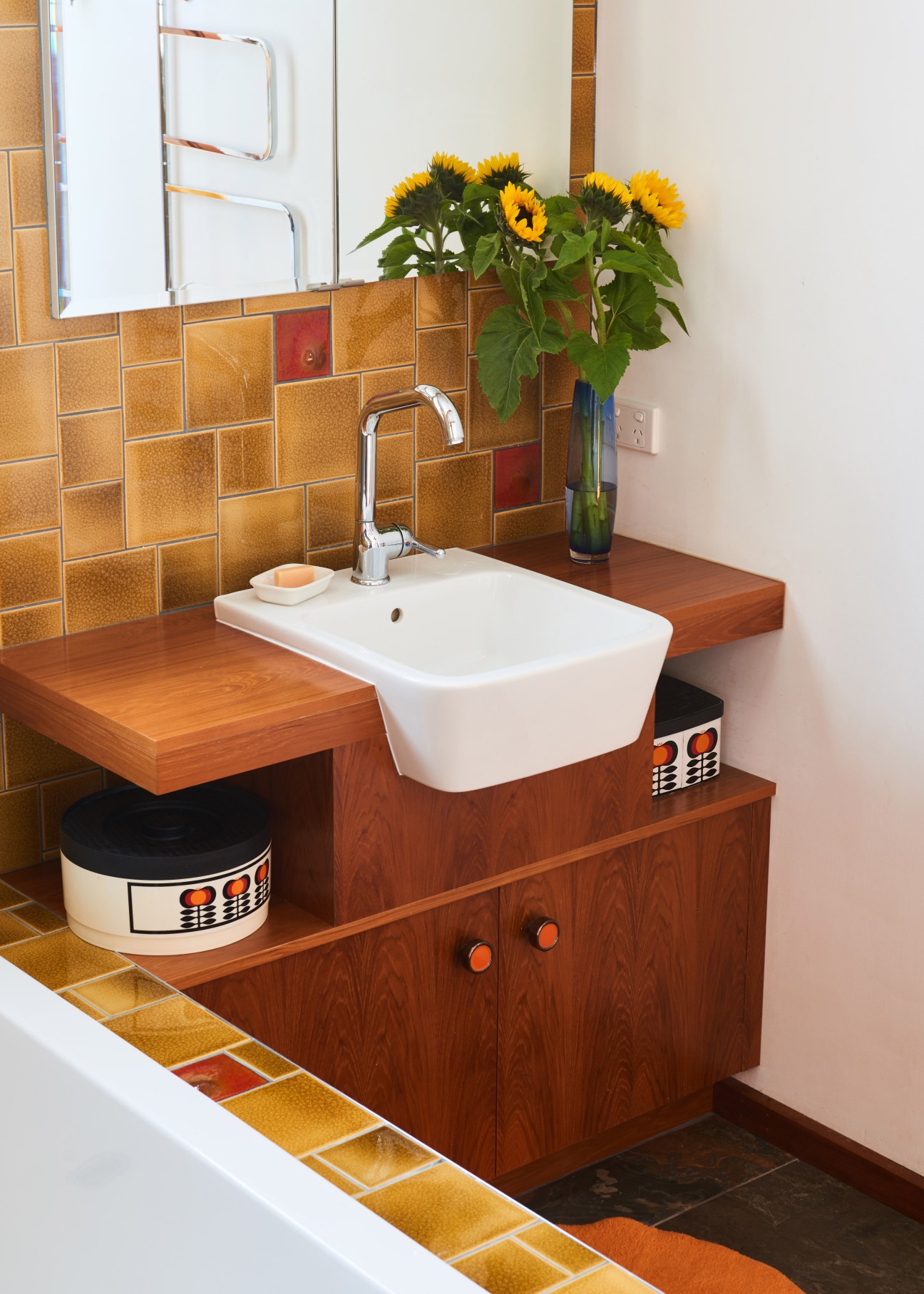 Wooden bathroom vanity with white sink, chrome faucet, golden tile backsplash, and sunflowers in vase.