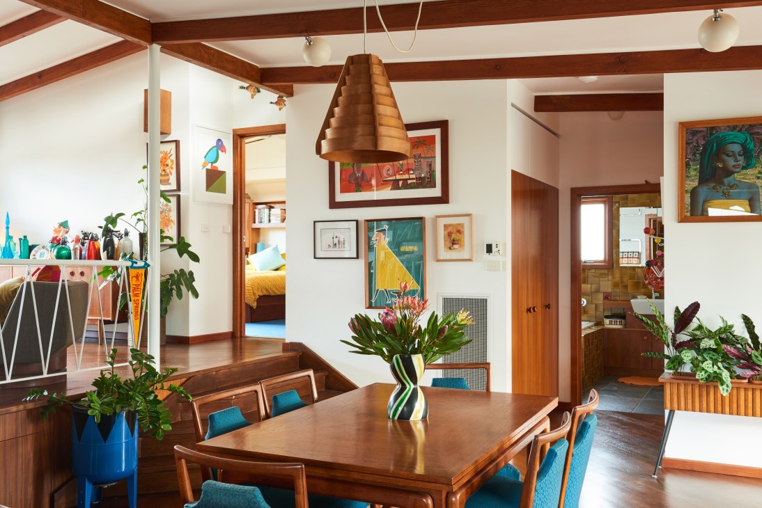 Mid-century modern dining and living area with exposed wooden beams, wood table, blue chairs, copper pendant light and colorful artwork on walls.