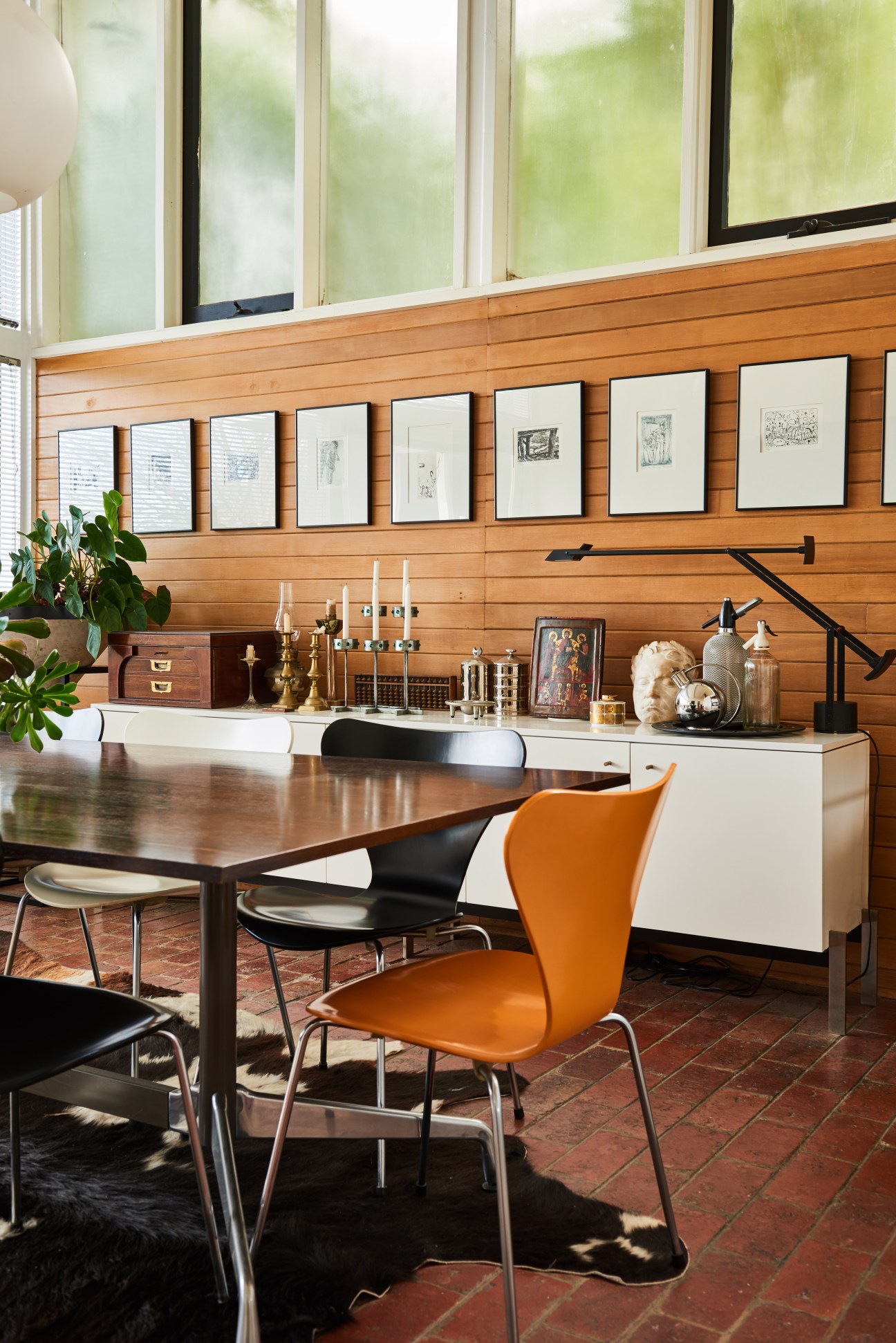 Modernist dining room with cedar-panelled walls, framed artwork, orange chair, and table with decorative objects.