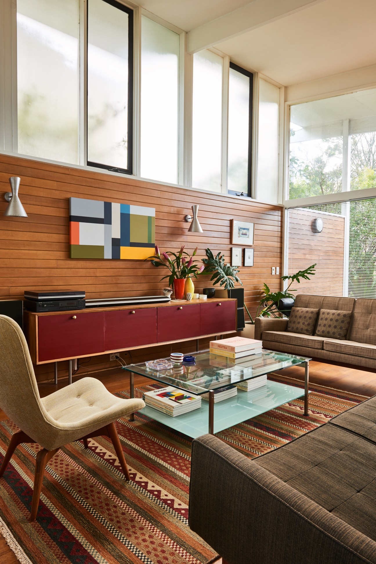 Mid-century modern lounge with wooden paneling, red credenza, cream chair, and floor-to-ceiling windows overlooking trees.