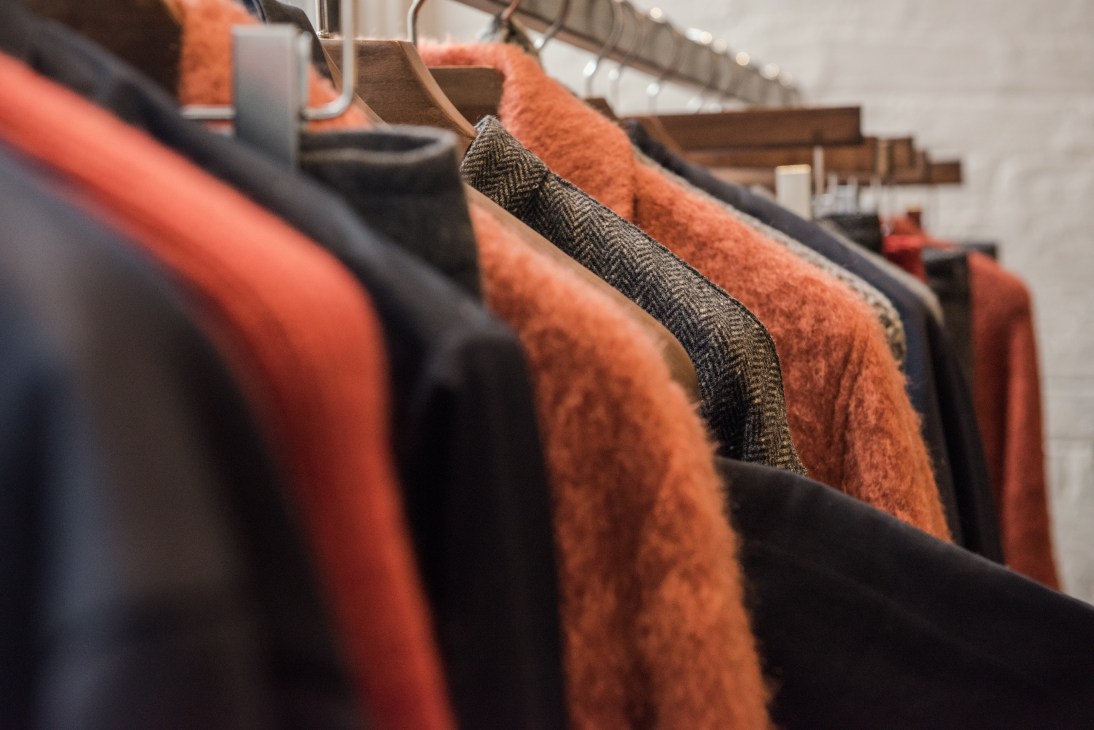 Deveaux clothing samples on wooden hangers, including burnt-orange brushed wool and dark herringbone fabrics.
