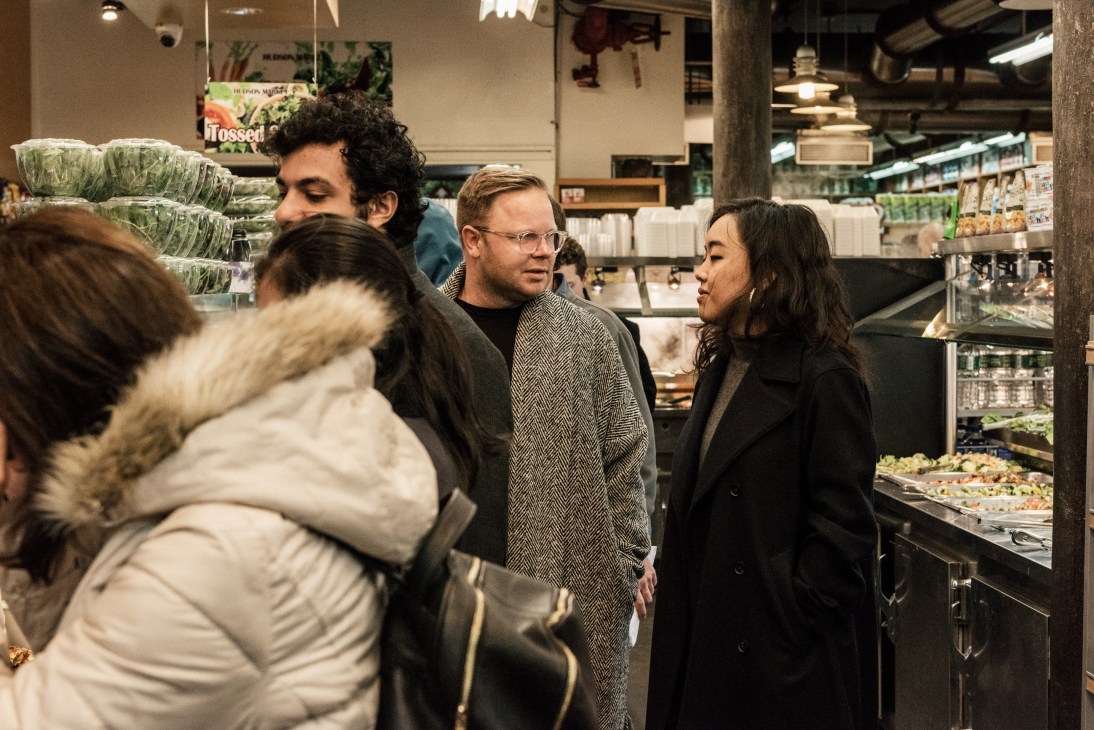 Matthew Breen and Andrea Tsao stand in Hudson Market bodega wearing Deveaux pieces amid other customers and prepared food displays.