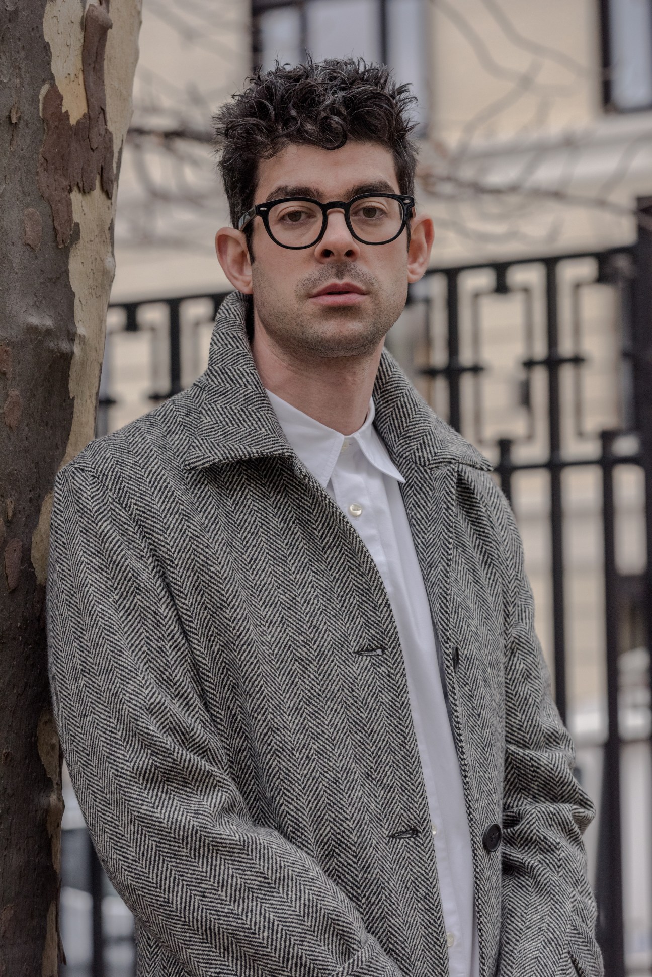 Man wearing black-framed glasses and grey herringbone car coat over white shirt, leaning against weathered concrete wall.