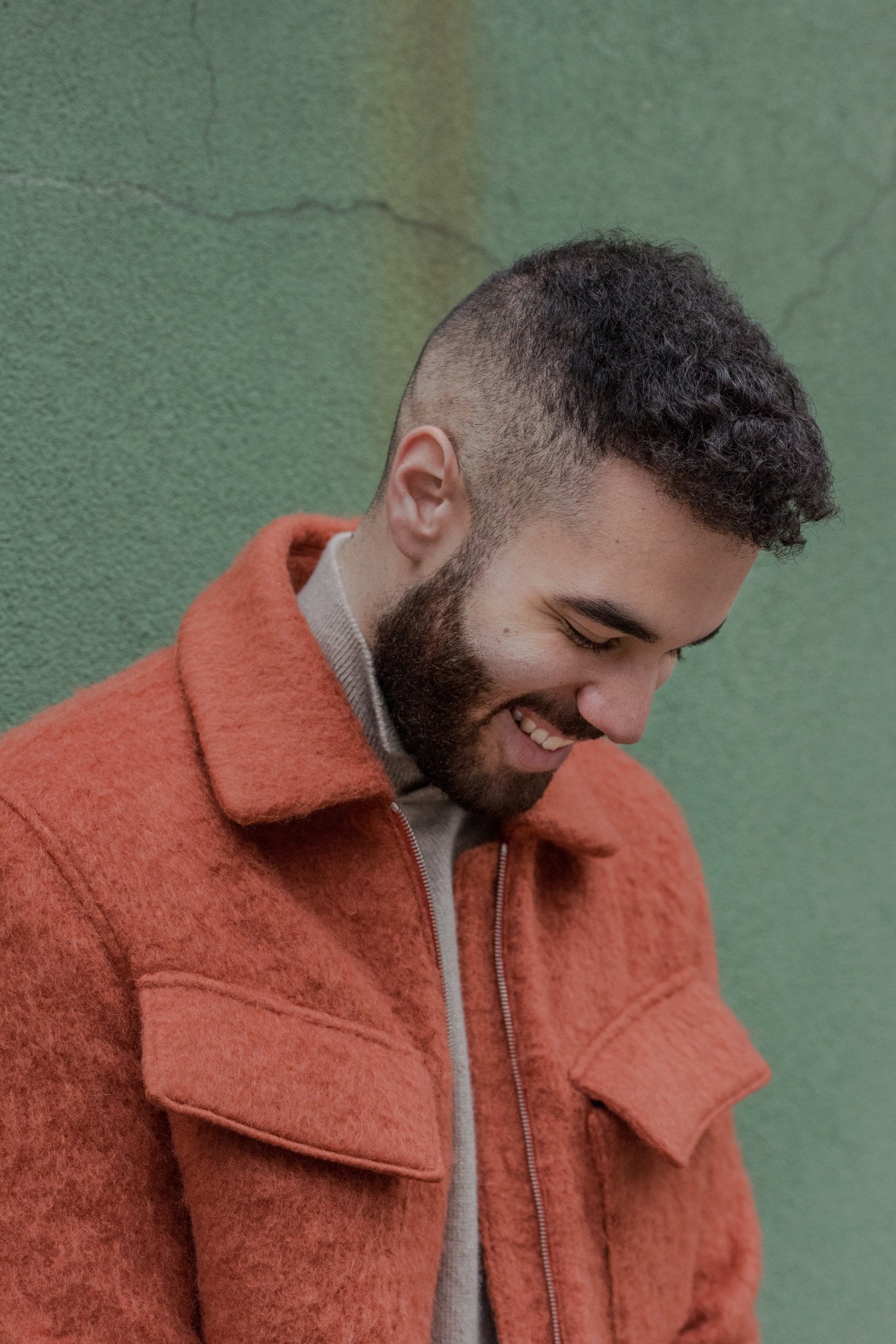 Man wearing Deveaux split neck sweater and brushed wool Harrington jacket in rust orange against green wall.