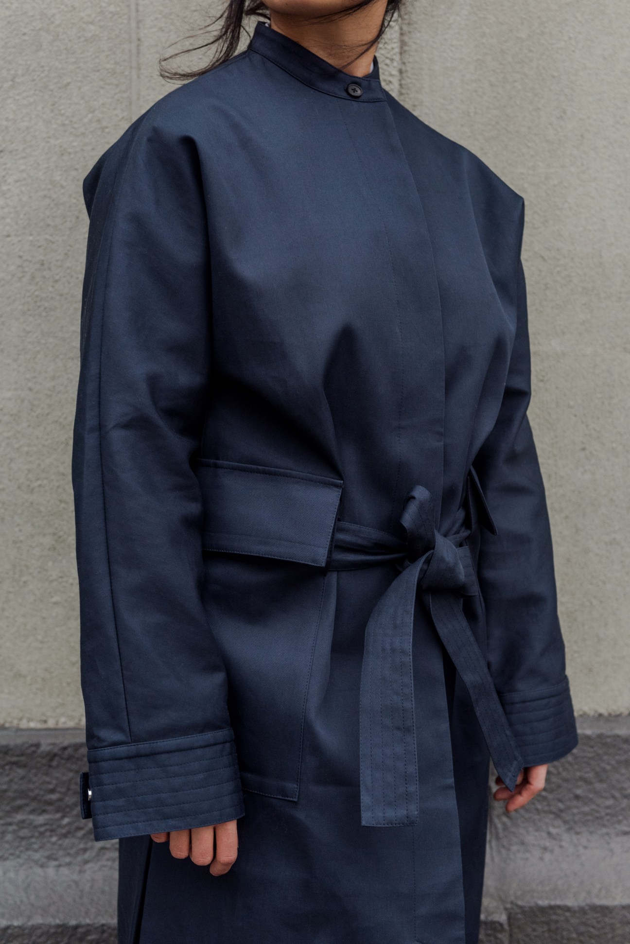 Woman wearing Deveaux navy coat dress with tied waist and high neckline against grey wall.