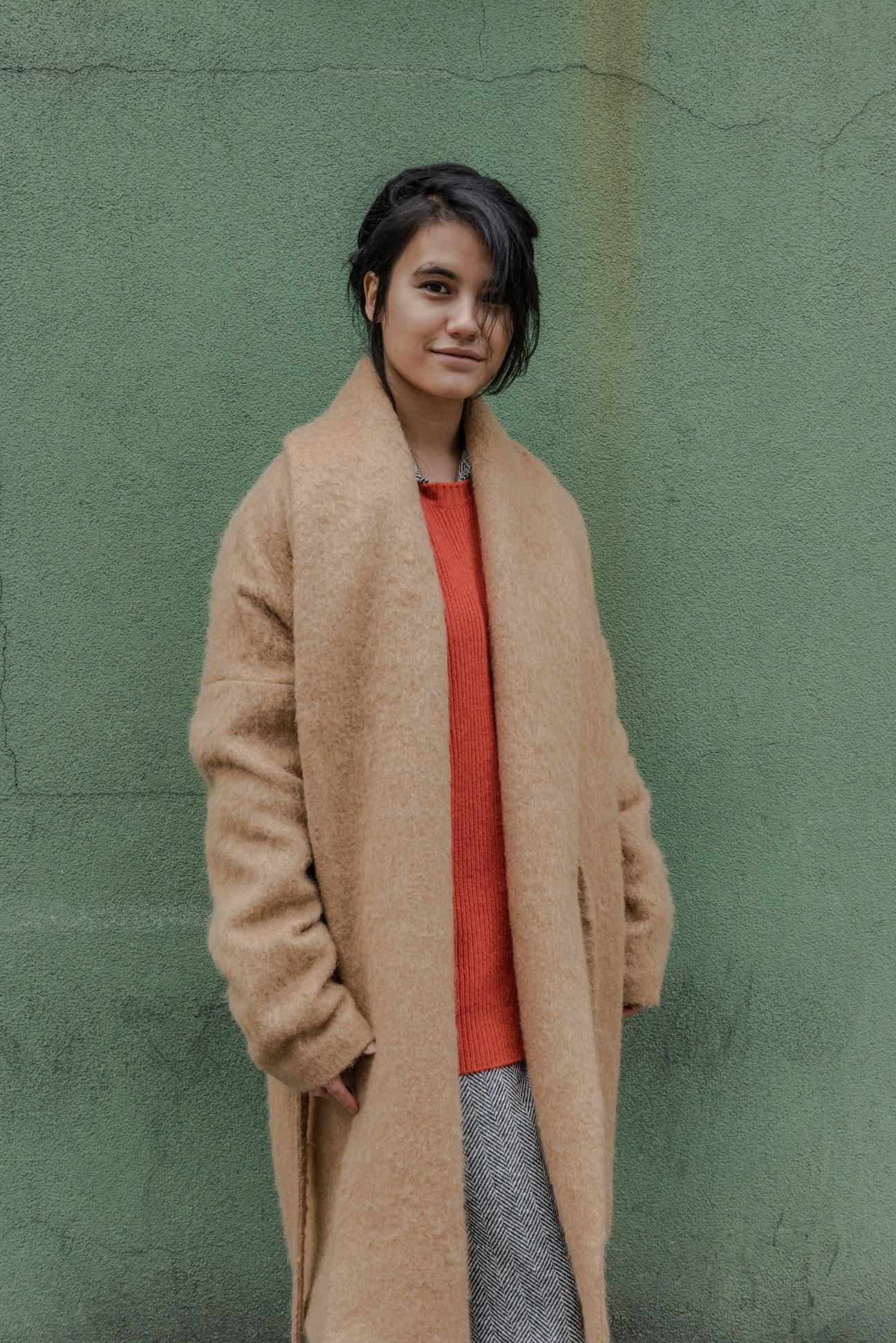Woman wearing a camel side-tie coat over an orange ribbed cashmere sweater against a green wall.