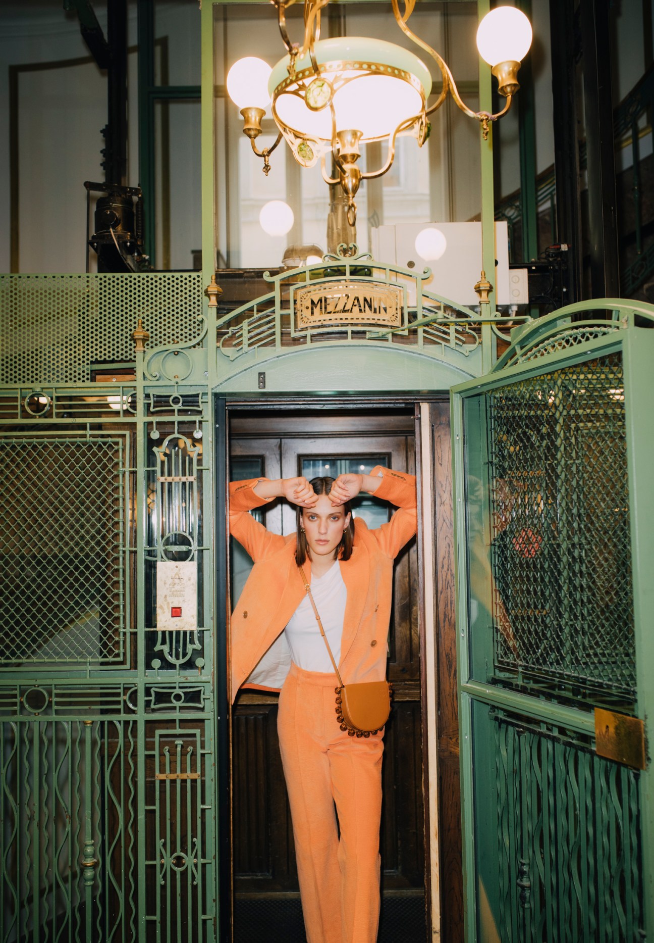 Woman in orange suit standing in ornate vintage elevator with brass fixtures and green metalwork