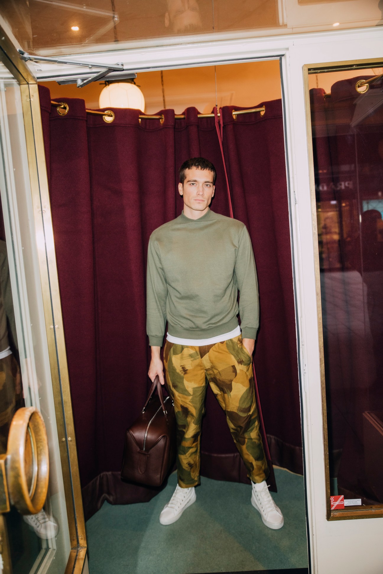 Man in olive sweatshirt and camouflage trousers standing in train compartment with leather bag