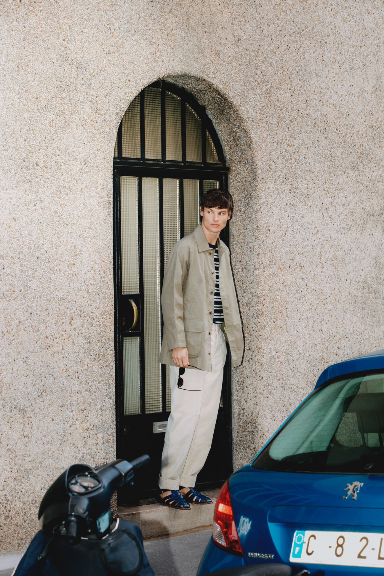 Young man in beige jacket and striped shirt standing in arched doorway next to blue car