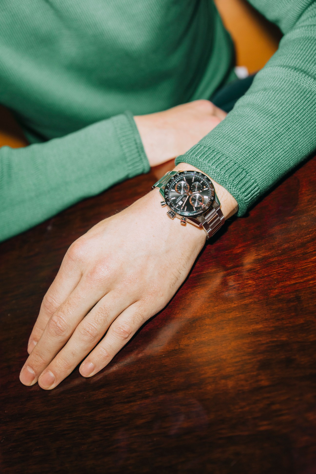 Hand wearing a Tag Heuer chronograph watch and green Altea jumper resting on wooden table