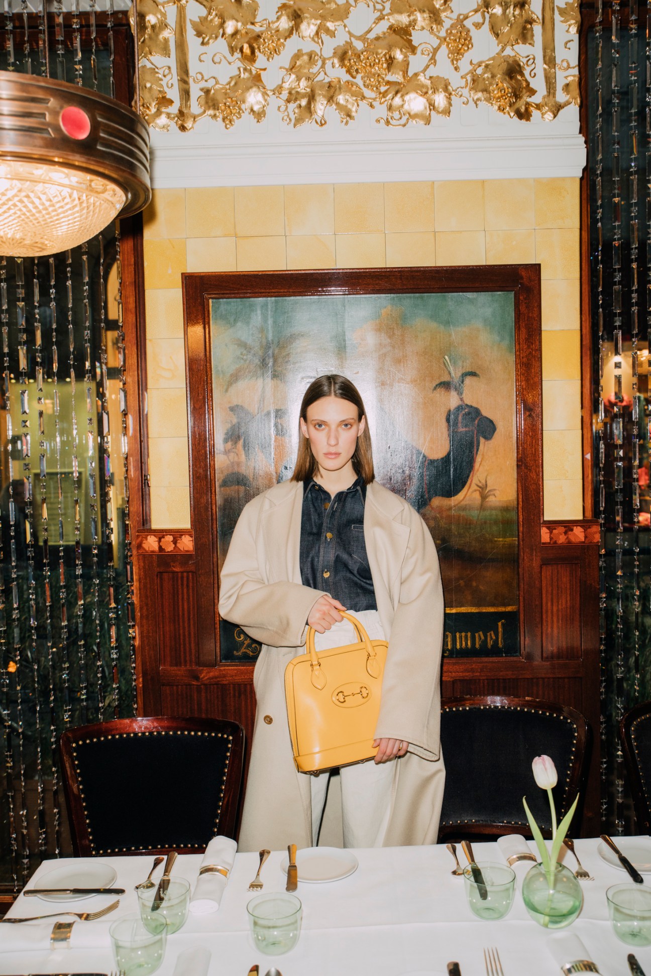 Woman in beige coat holding yellow handbag standing in ornate restaurant with decorative ceiling and set table