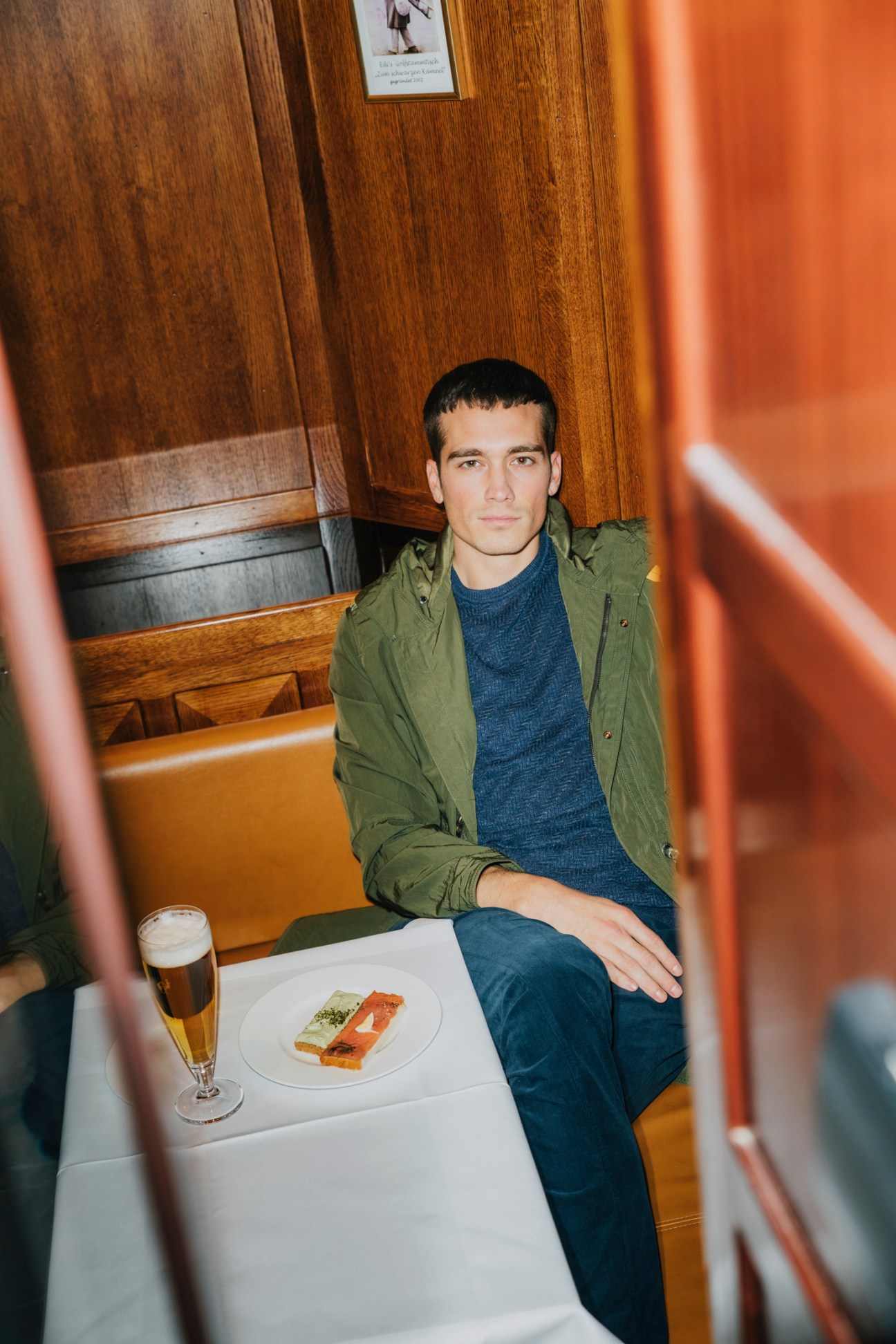 Young man in olive green coat and blue sweater sitting at cafe table with beer and food plate