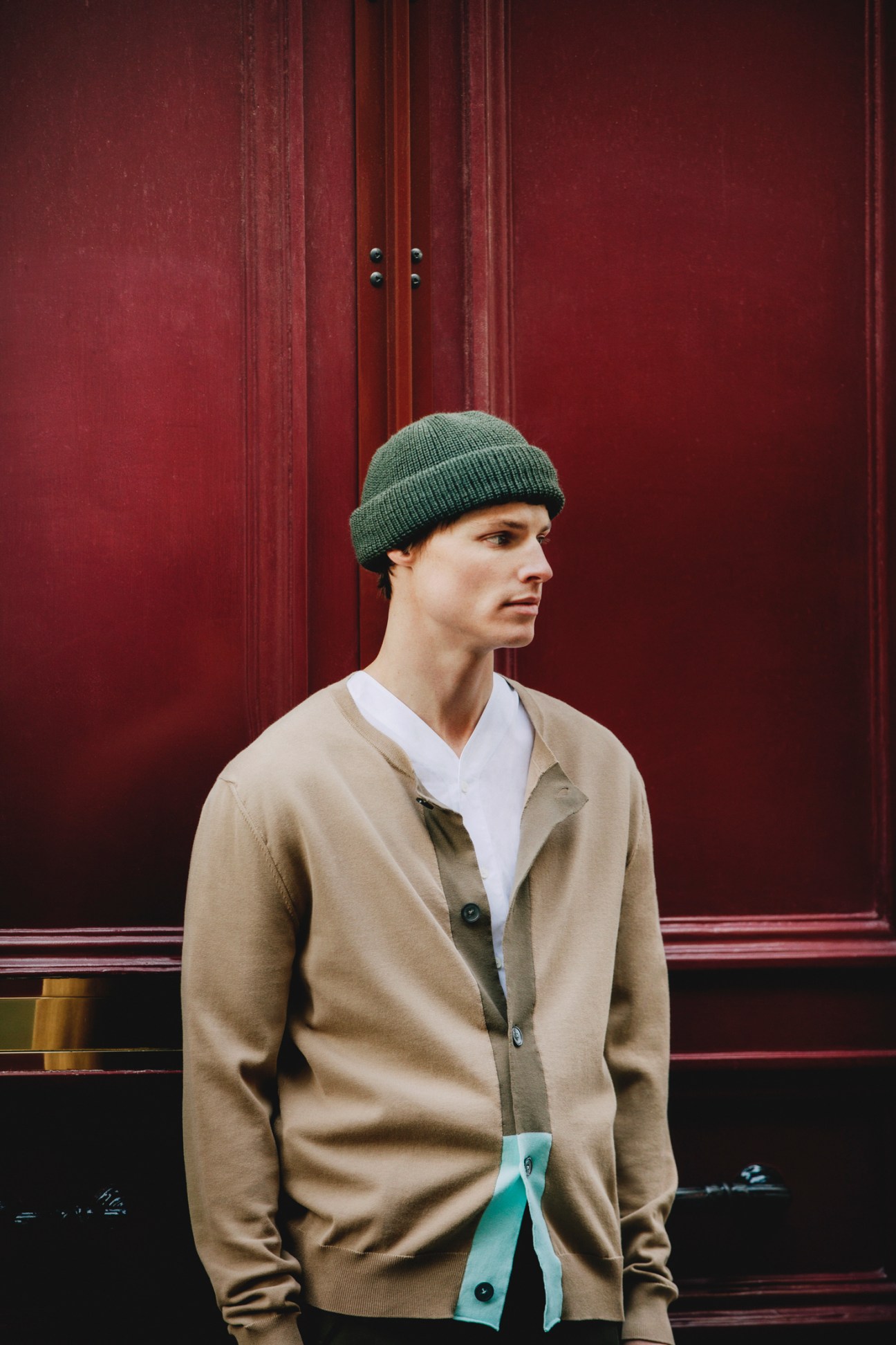 Man in beige cardigan and green beanie standing against red doors