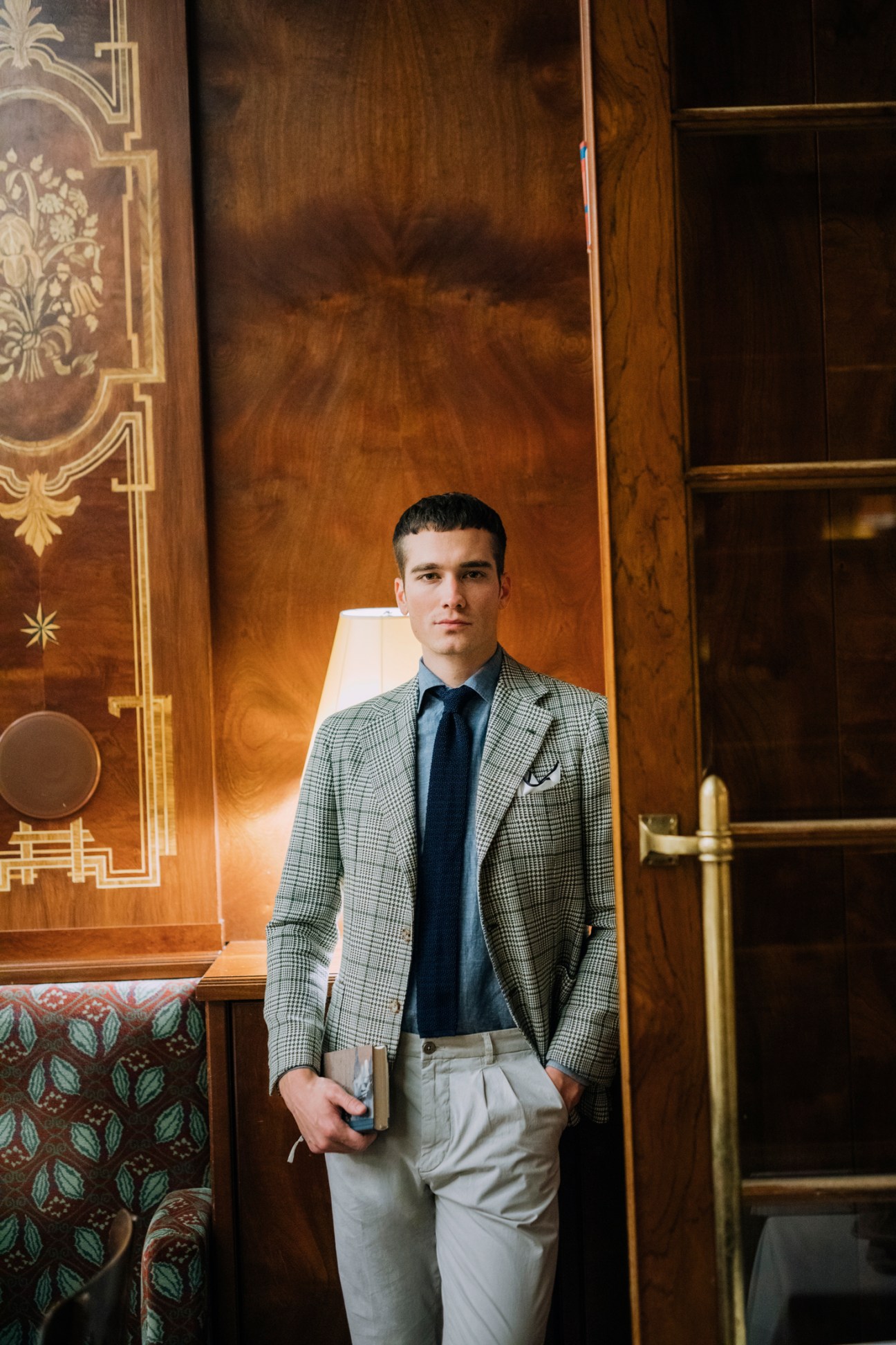 Man in grey checked jacket, navy tie and light trousers standing against ornate wooden paneling