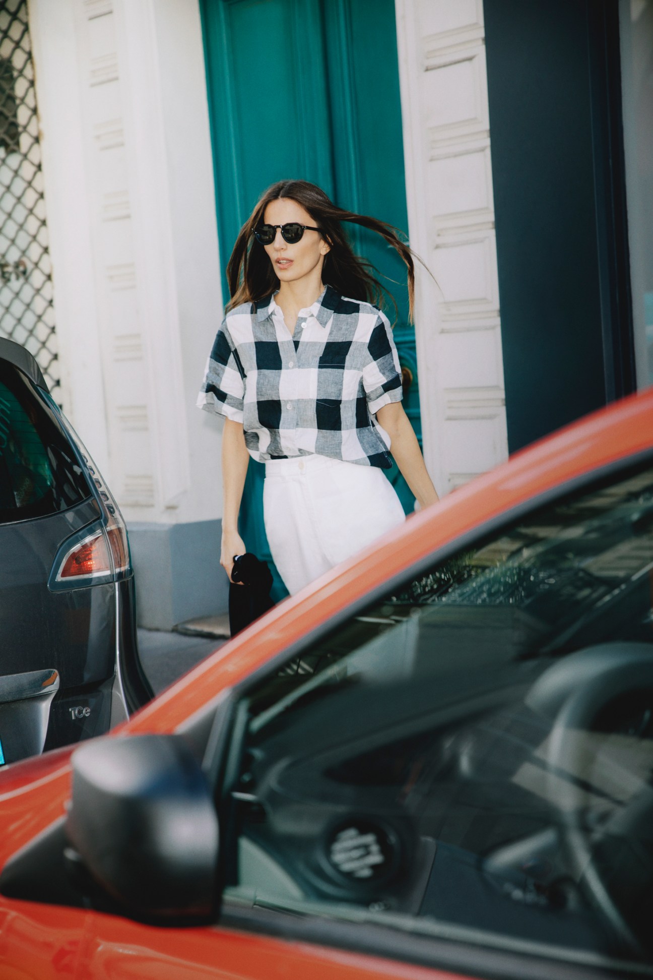 Woman in black and white checkered shirt and white trousers walking between parked cars on city street