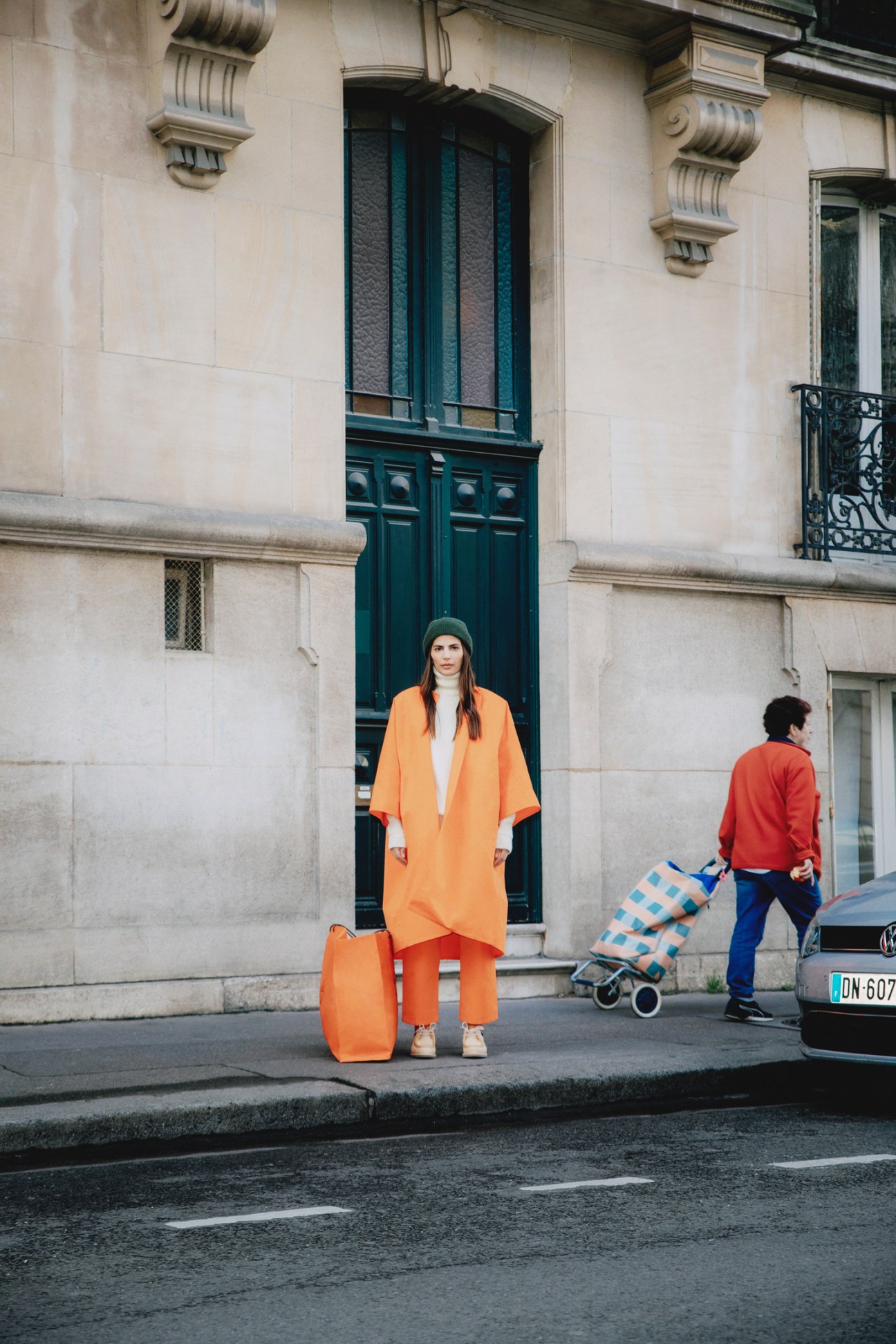 Woman in orange suit and green beanie stands on Parisian street with classical architecture