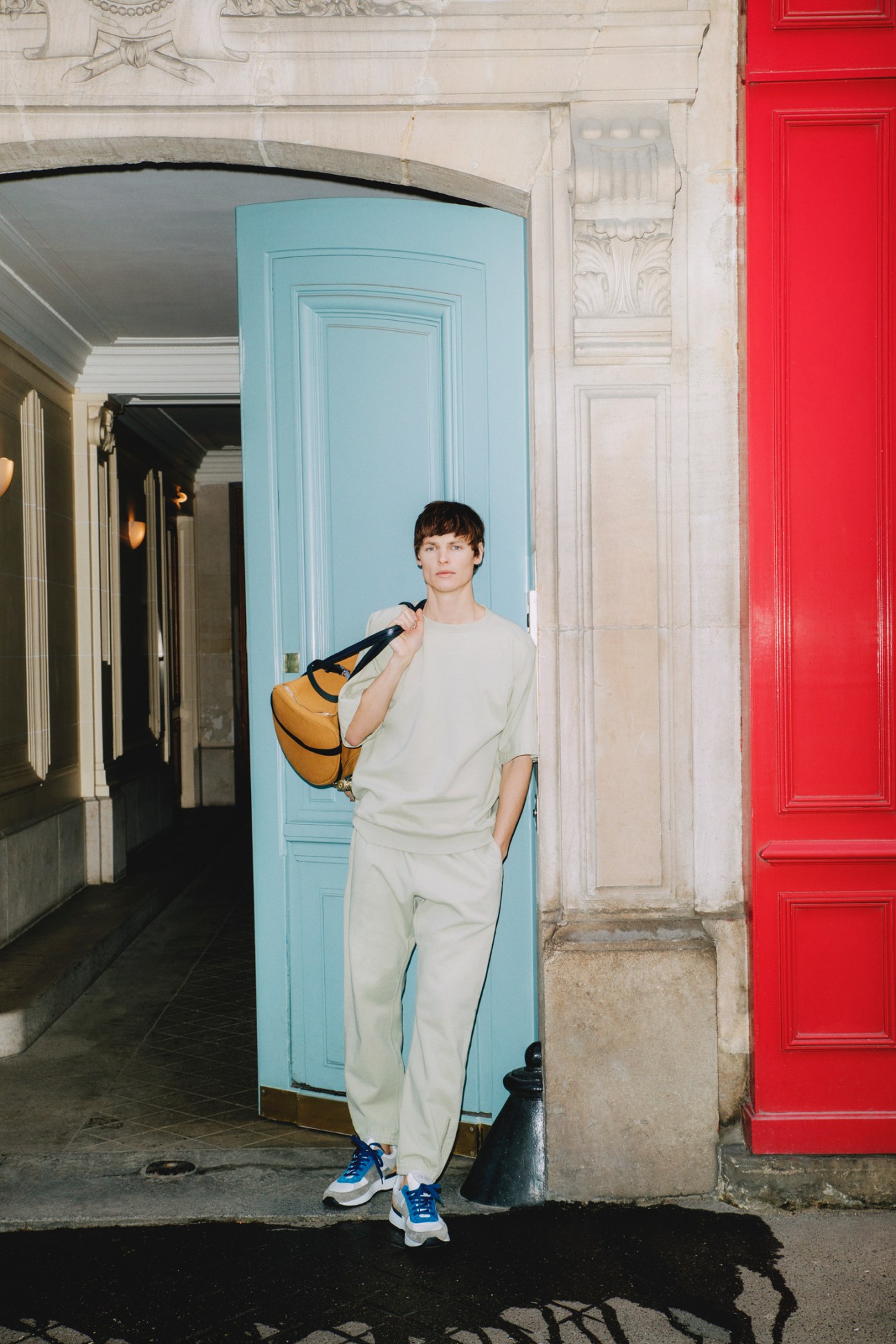 Young man in cream sweatsuit carrying yellow bag stands in ornate Parisian doorway with colorful doors
