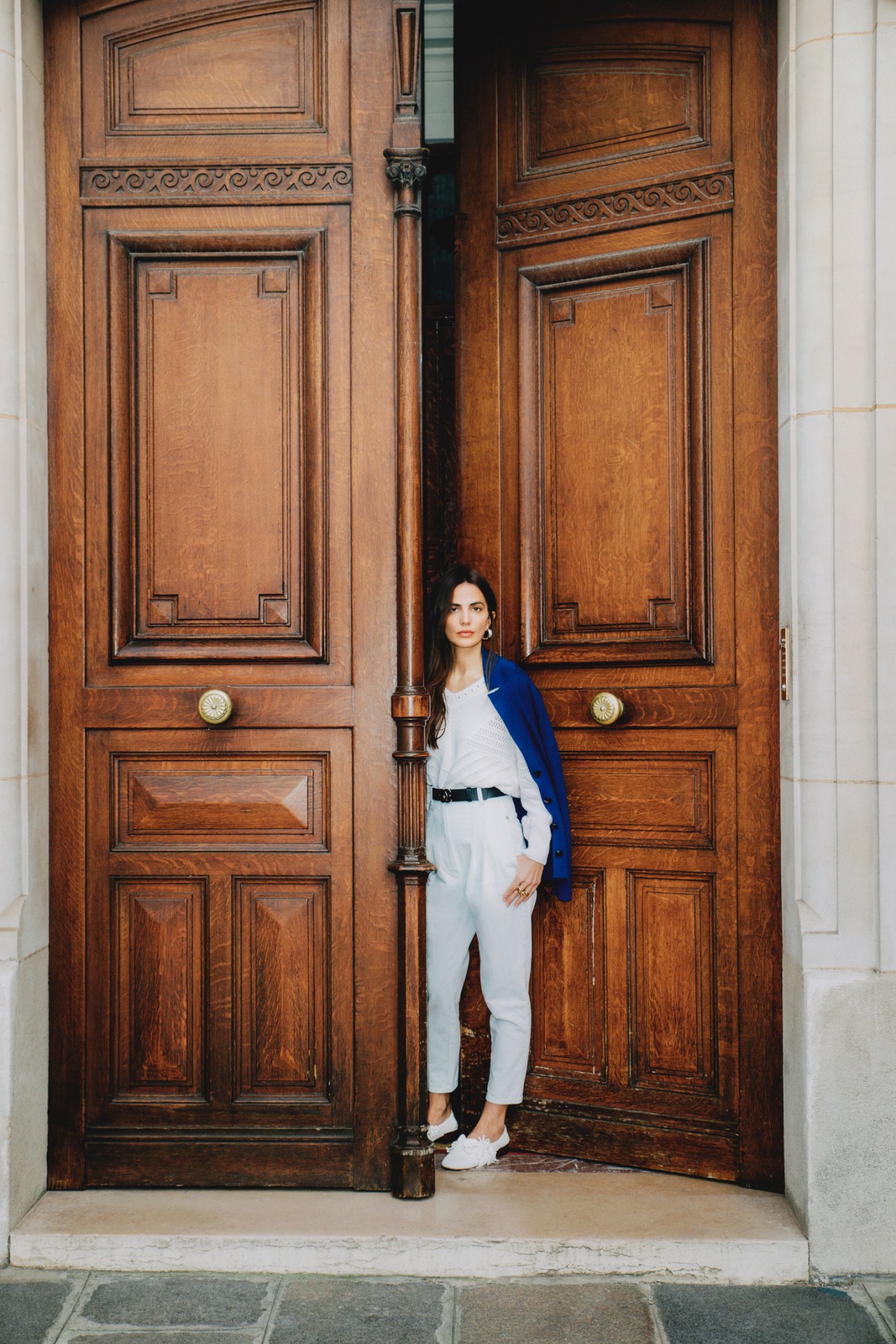 Woman in white outfit with blue jacket standing against ornate wooden doors in Paris