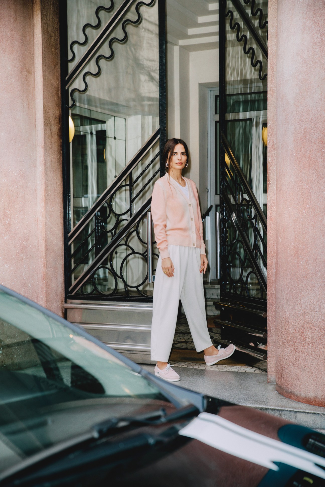 Woman in pink cardigan and white trousers standing on steps outside glass doors with ornate ironwork