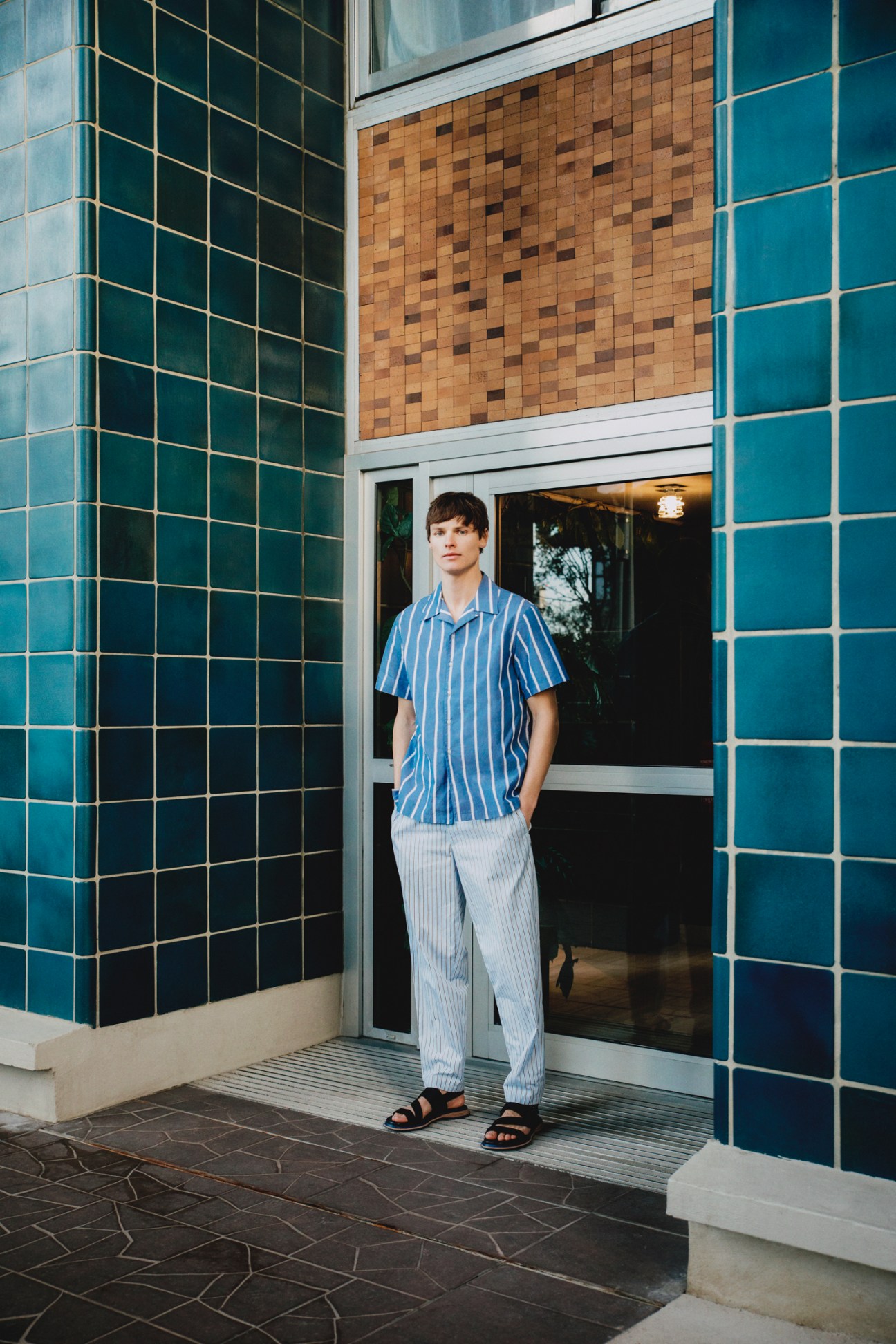 Young man in blue striped shirt and light trousers standing in doorway of tiled building