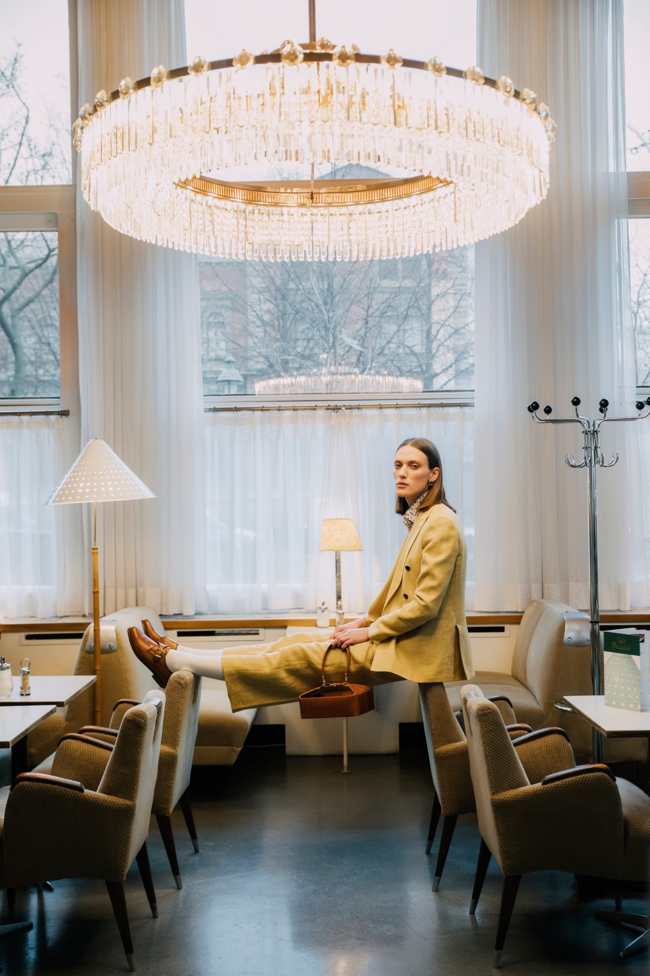 Woman in yellow suit sitting in elegant cafe interior with crystal chandelier and large windows