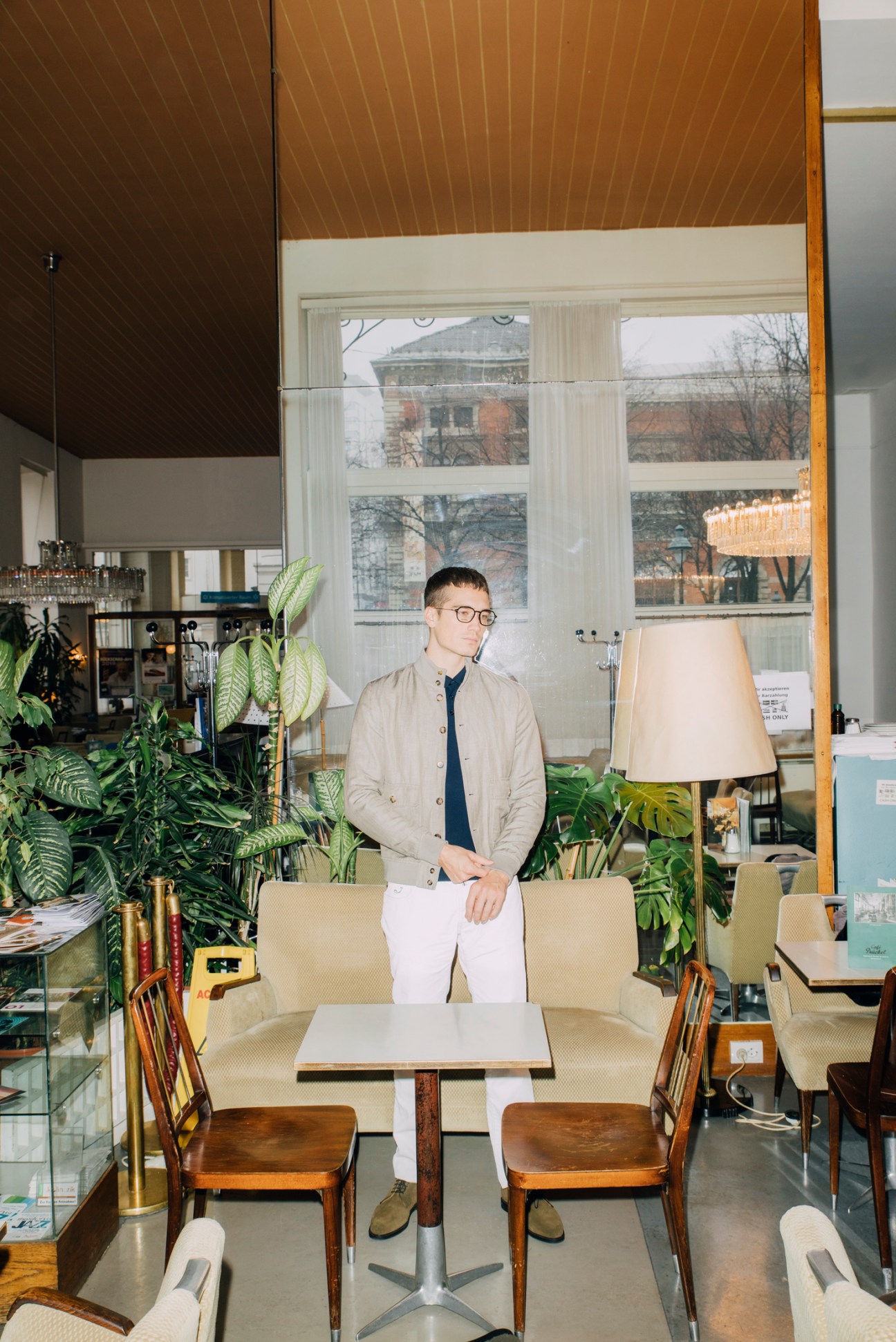 Man in glasses wearing beige jacket and dark polo shirt sitting in modern cafe with plants and large windows