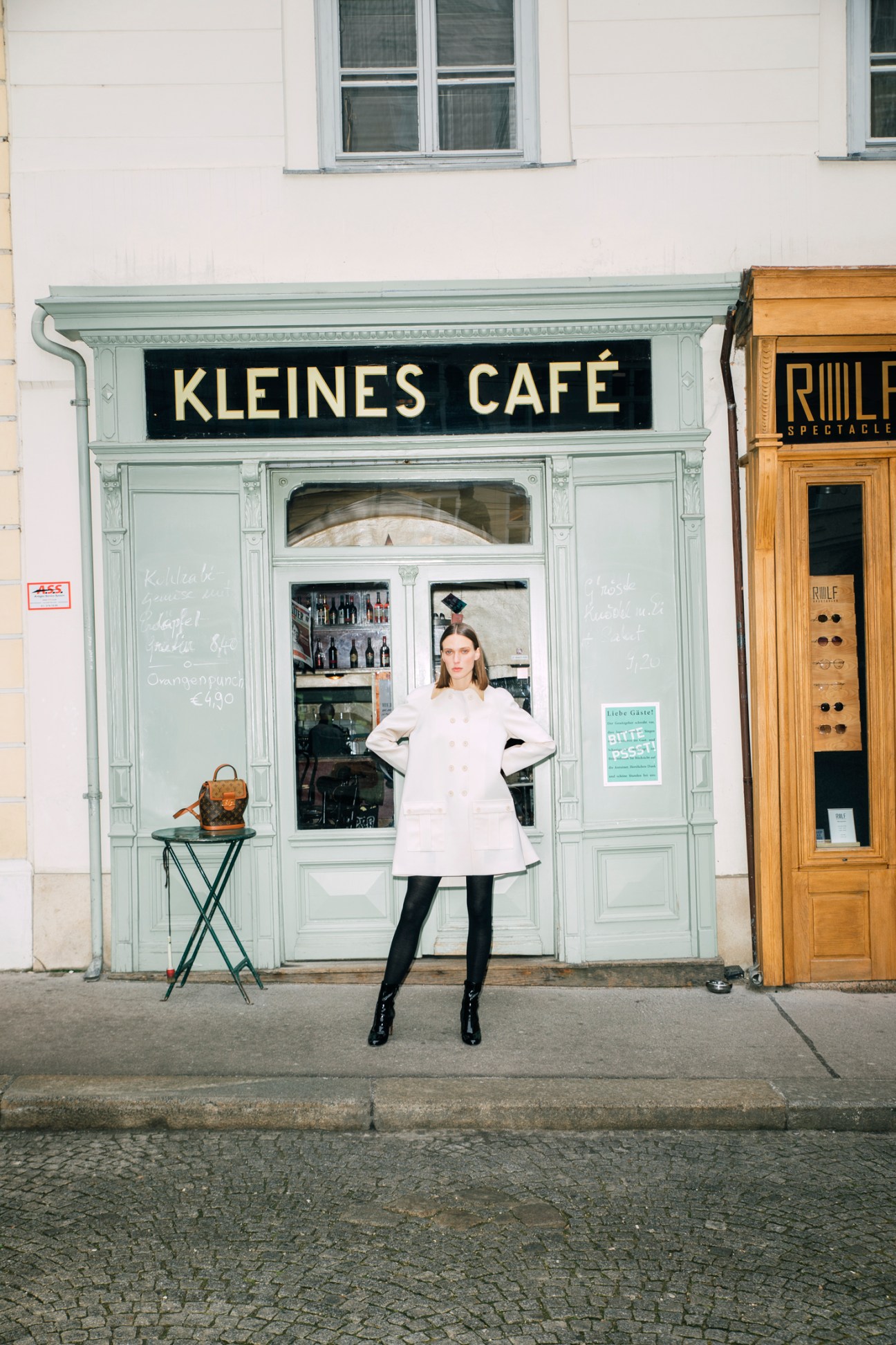Woman in white Louis Vuitton coat and boots standing in front of Kleines Café storefront