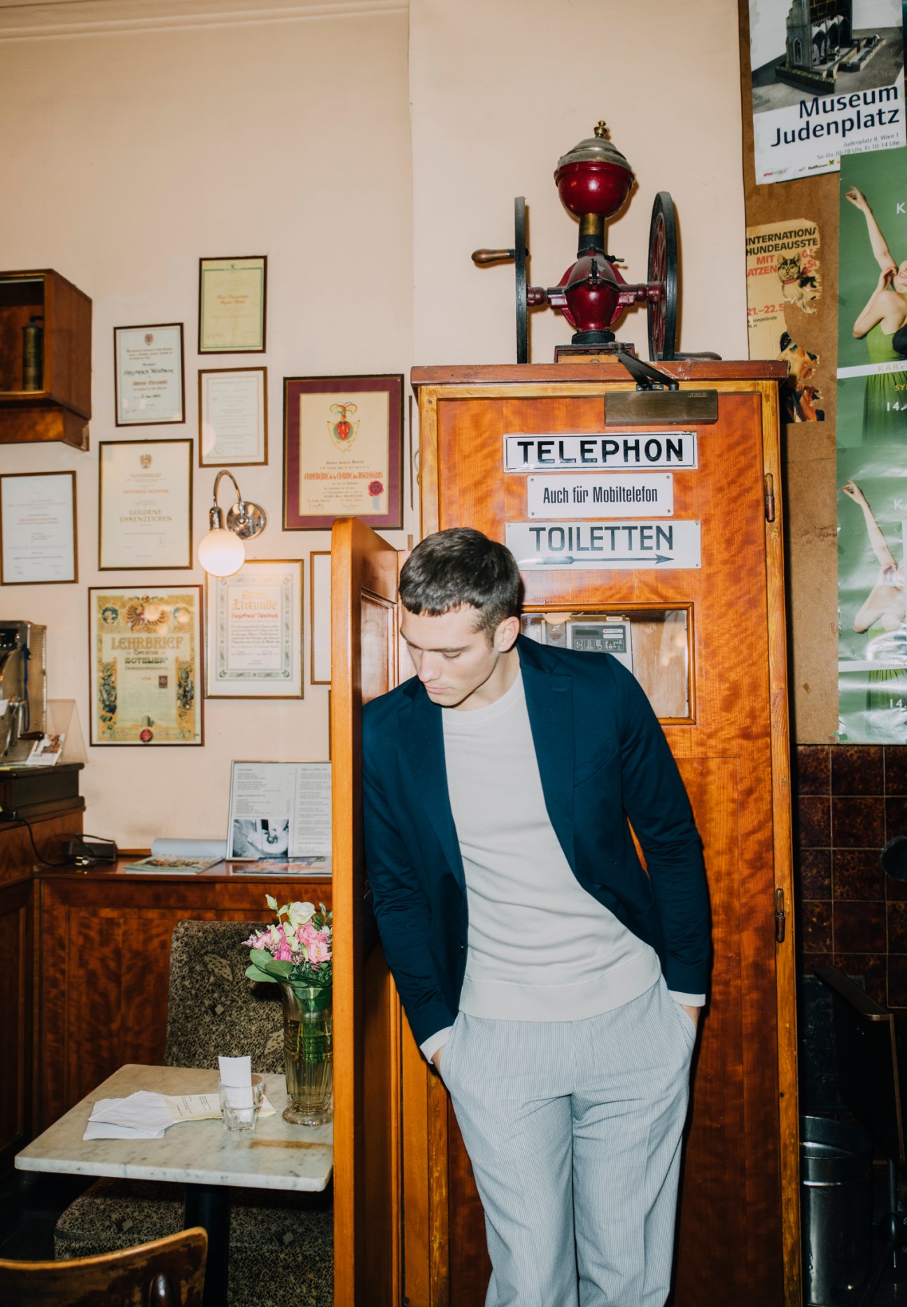 Man in dark jacket and light trousers leaning against vintage wooden telephone booth in traditional Austrian cafe