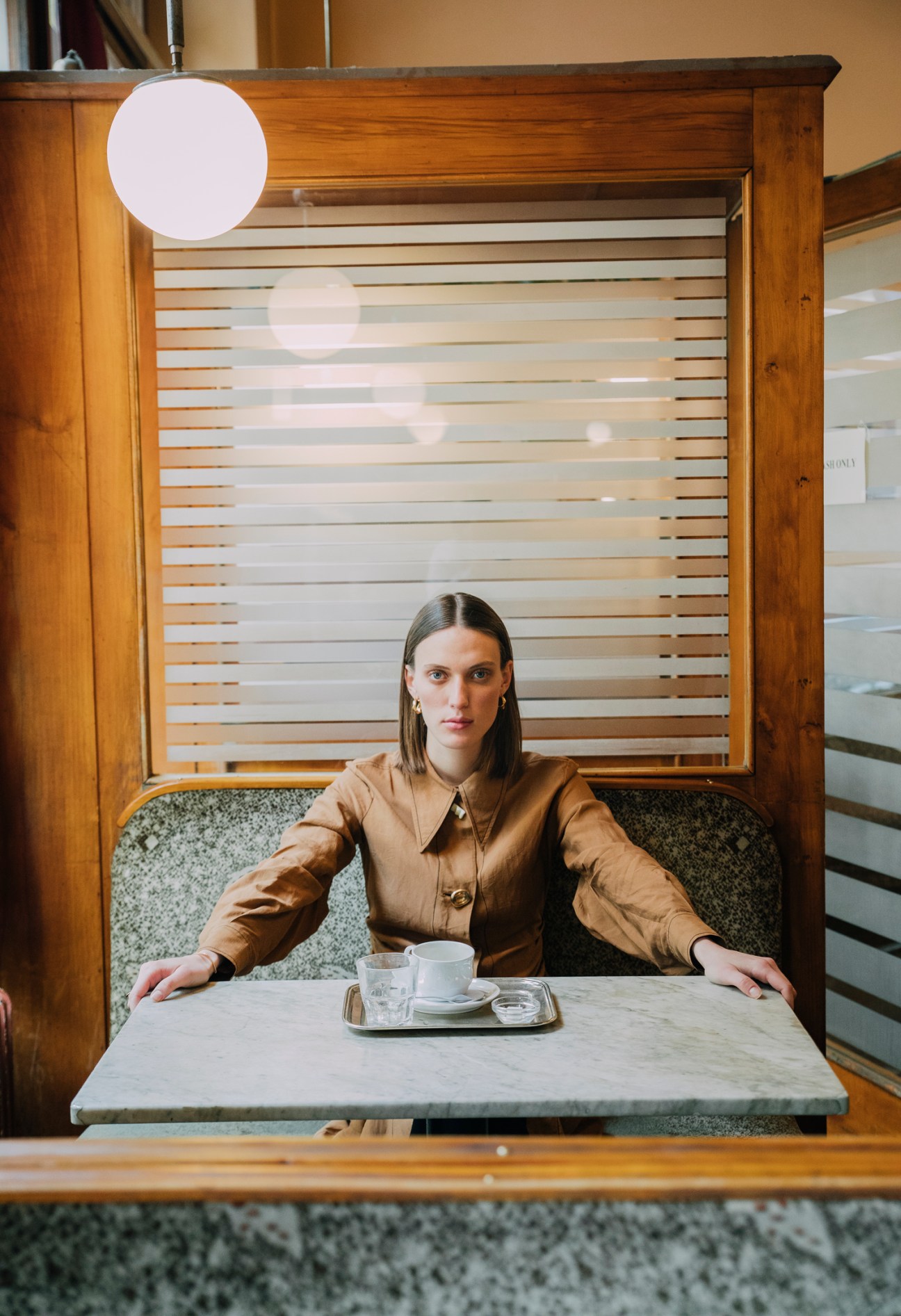 Woman in beige dress sitting at marble cafe table with coffee cup in wooden booth