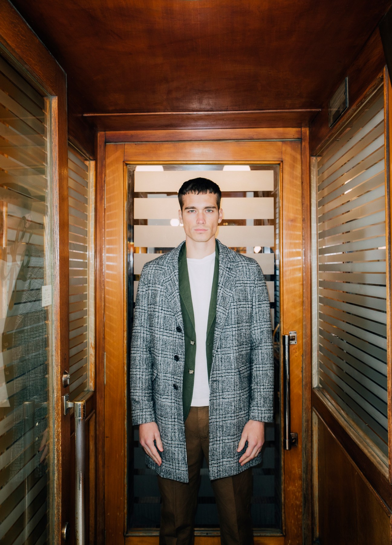 Man in grey tweed coat and green cardigan standing in wooden doorway