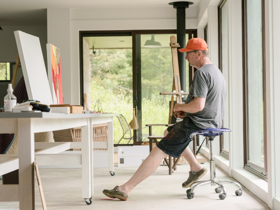 Artist Gary Hume sitting on a stool in his bright studio, viewing a canvas on an easel with large windows behind him.