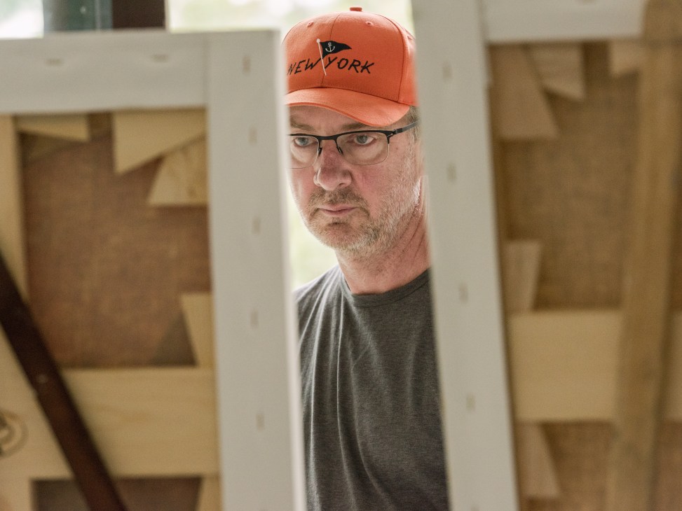 Artist Gary Hume wearing a red New York cap and glasses, peering through a white easel in his studio.