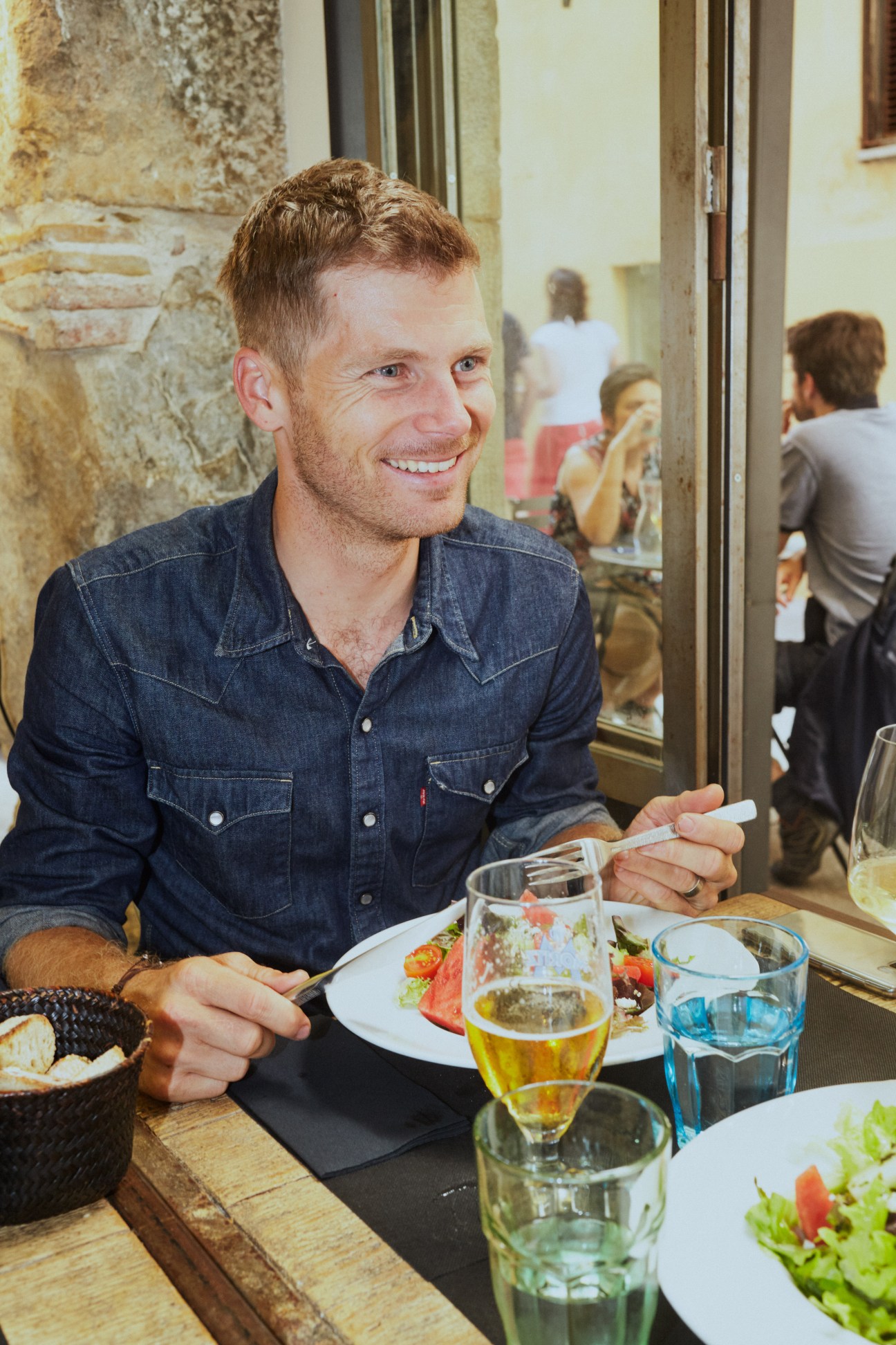 A smiling man in a denim shirt sitting at a restaurant table with food and drinks