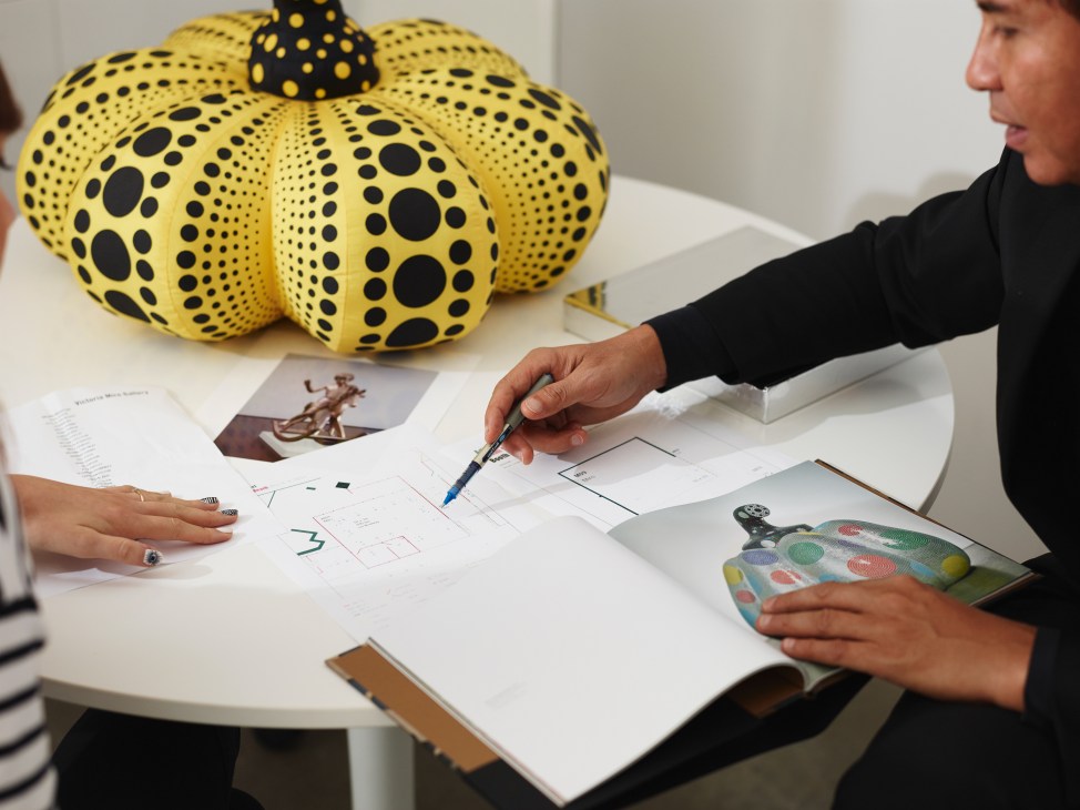 Gallery staff member discusses artwork and design concepts at a table with yellow polka-dotted sculpture and sketches.