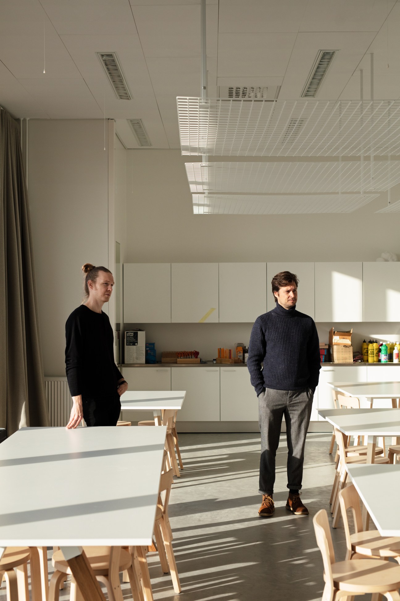 Two architects stand in a bright, minimalist kitchen space with wooden chairs and white cabinetry.