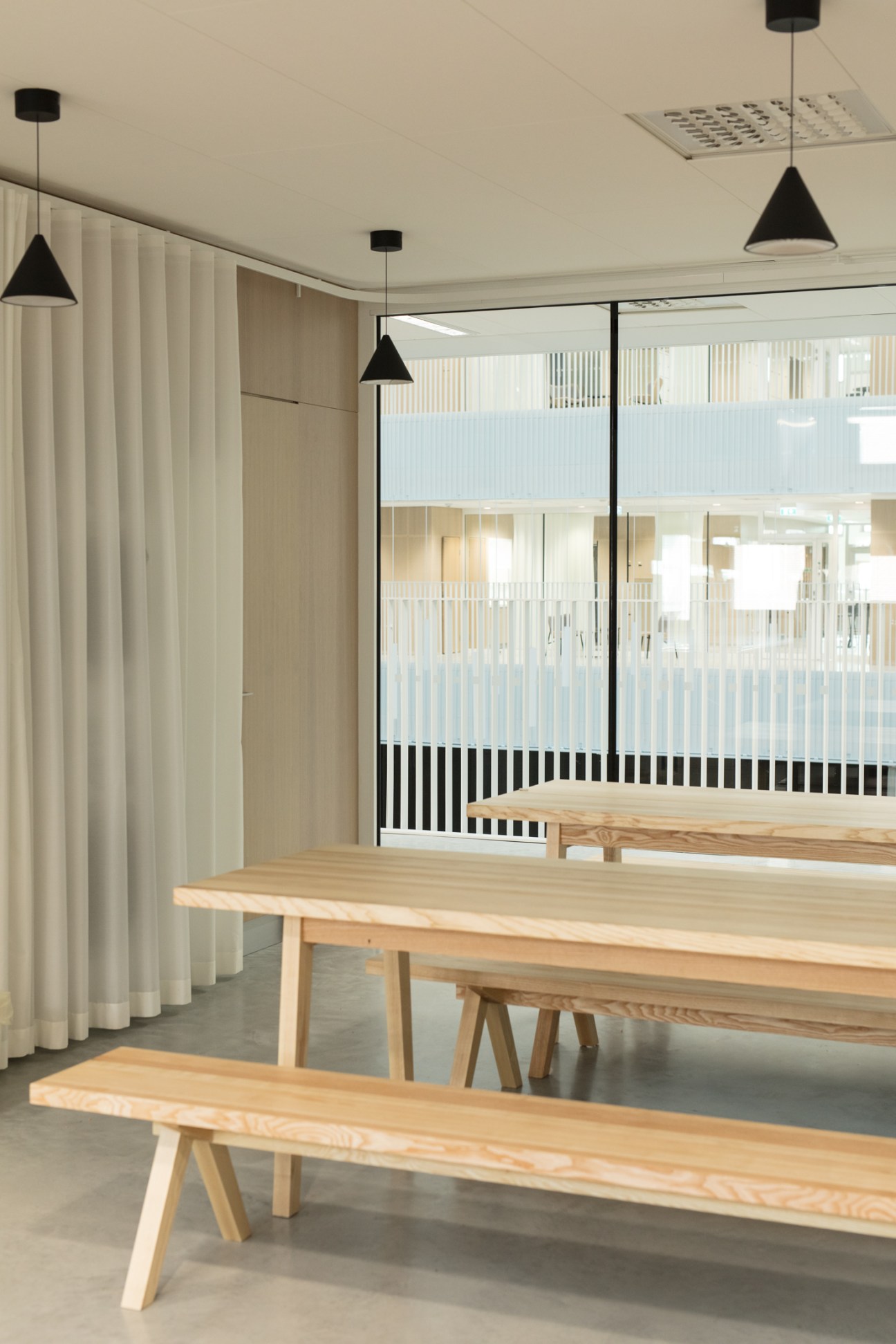 Minimalist classroom interior with wooden tables, black pendant lights, and floor-to-ceiling windows overlooking a school atrium.