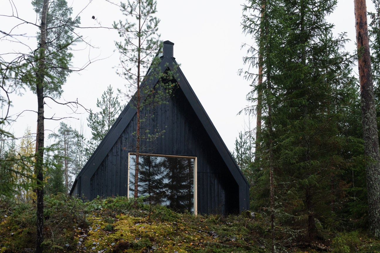 Black wooden chapel with pitched roof nestled among pine trees beside a lake in Finland.