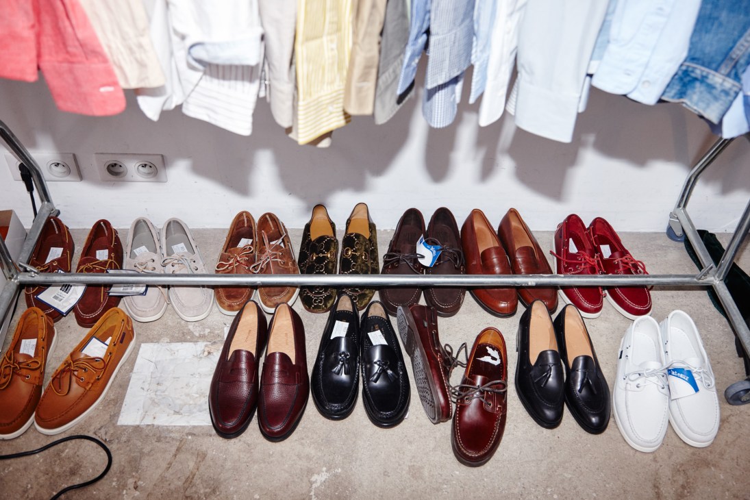 Selection of leather loafers, boat shoes and sneakers arranged on shelves with colorful shirts hanging above.