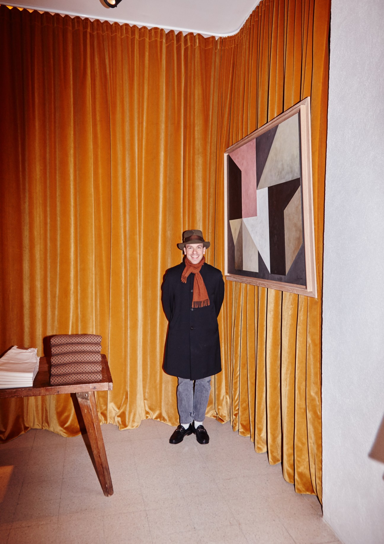 Gauthier Borsarello stands smiling in the Holiday Boileau shop wearing a navy coat, orange scarf and brown hat against golden curtains.