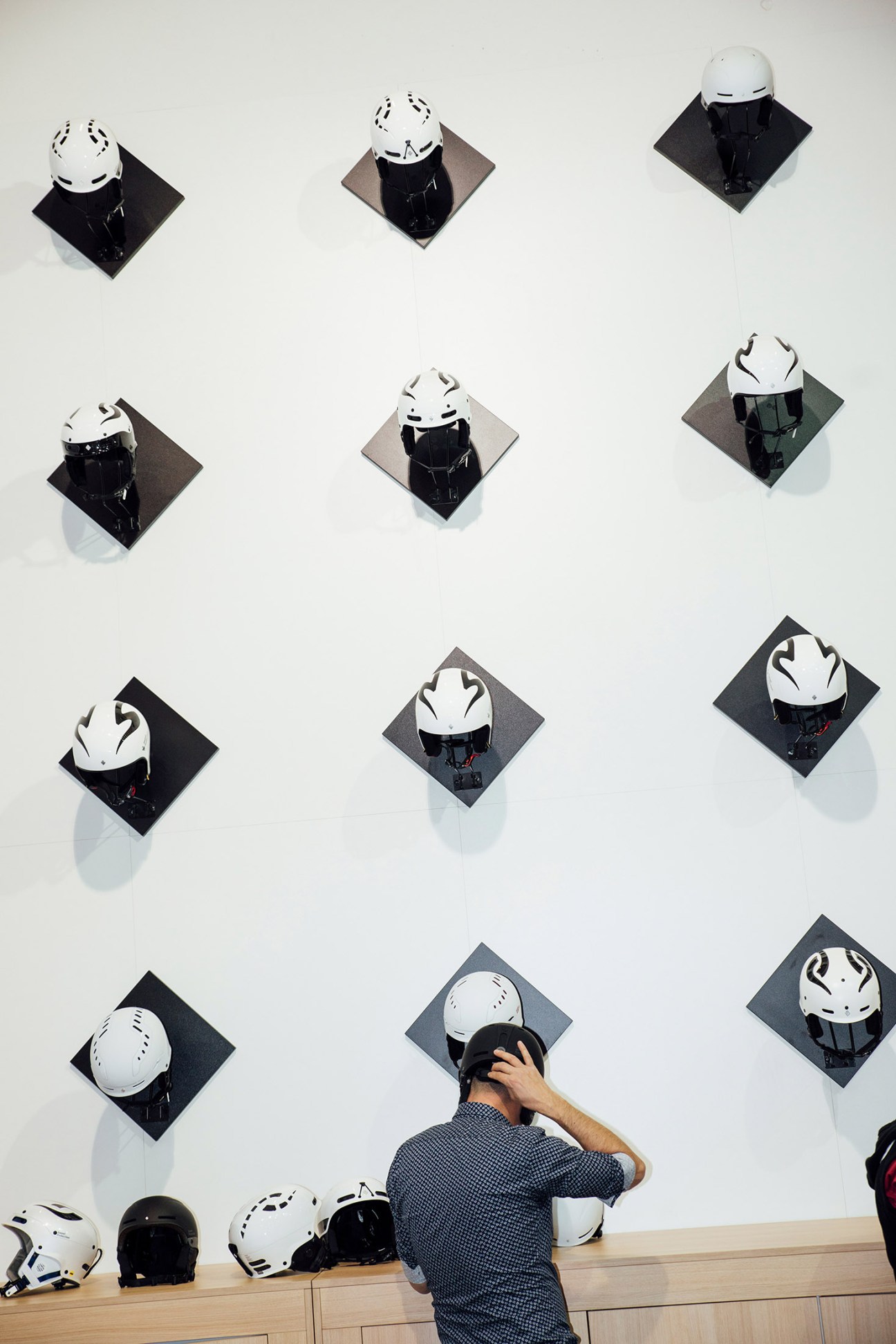 Visitor selecting a white ski helmet from a wall display of multiple helmets at Ispo sportswear fair.