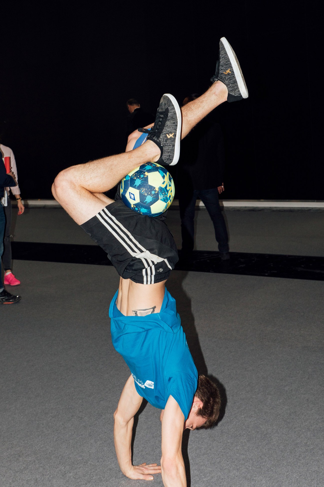 Person performing a handstand while balancing a soccer ball on their body at an indoor sports event.
