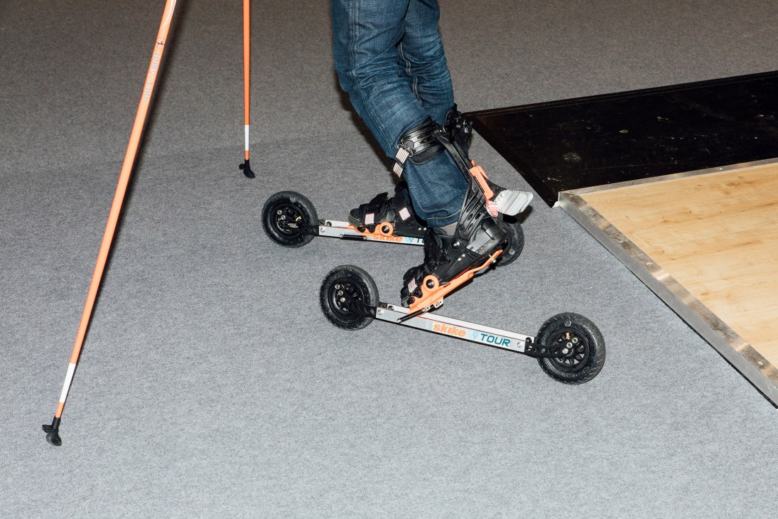 Person wearing jeans and orange and black ski roller skis on an indoor exhibition floor.