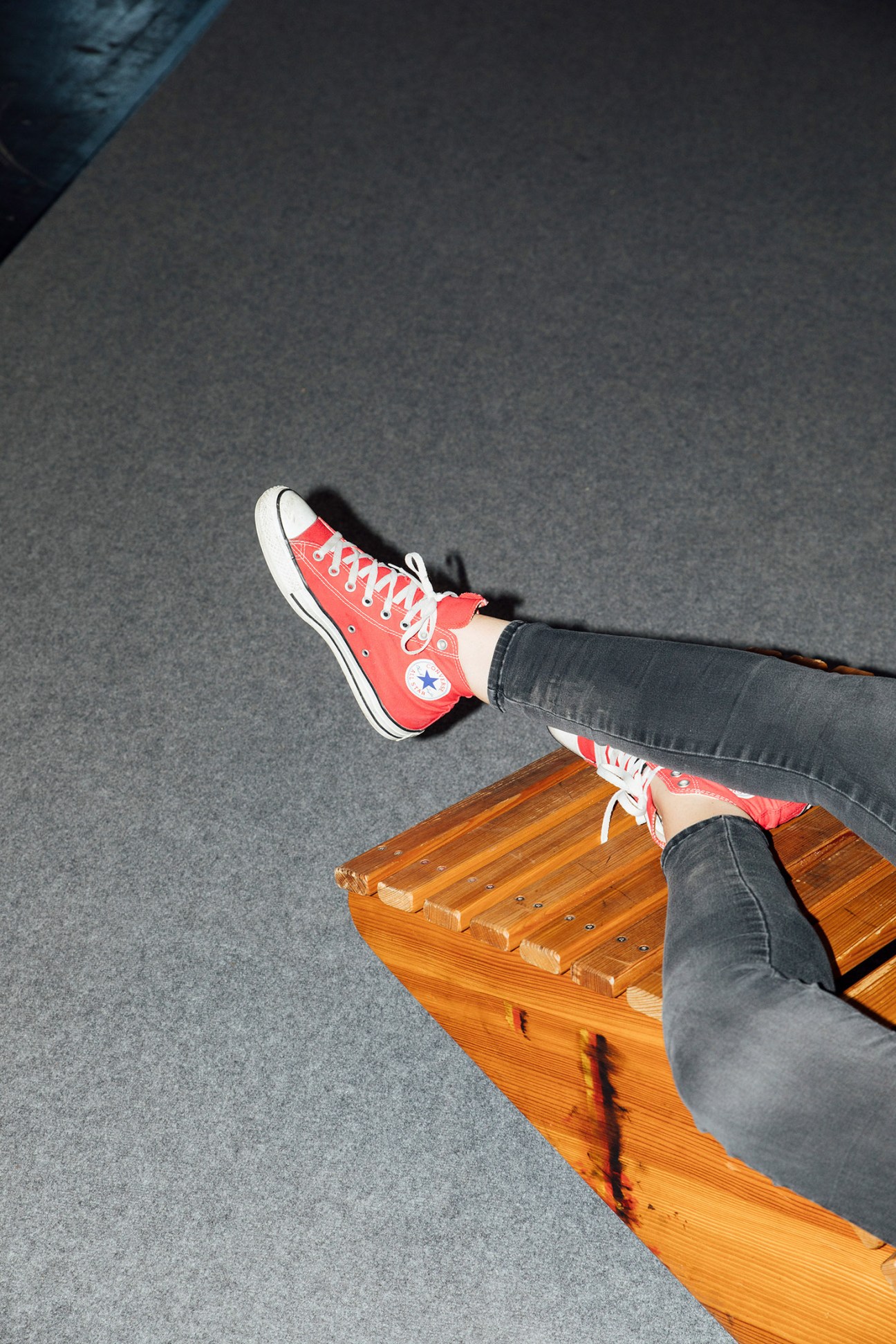 Person wearing red Converse high-tops with white laces, feet elevated on a wooden platform against grey flooring.