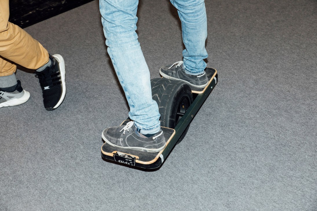 Person in jeans and sneakers riding an electric skateboard indoors.