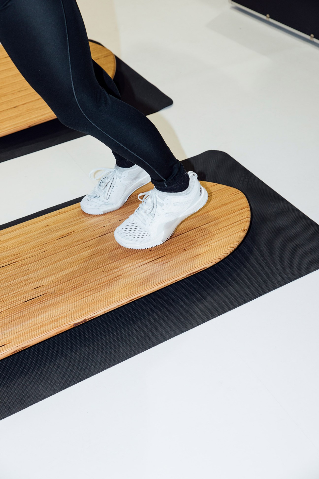 Person wearing white trainers and black leggings stretching legs on wooden floor with black mat.