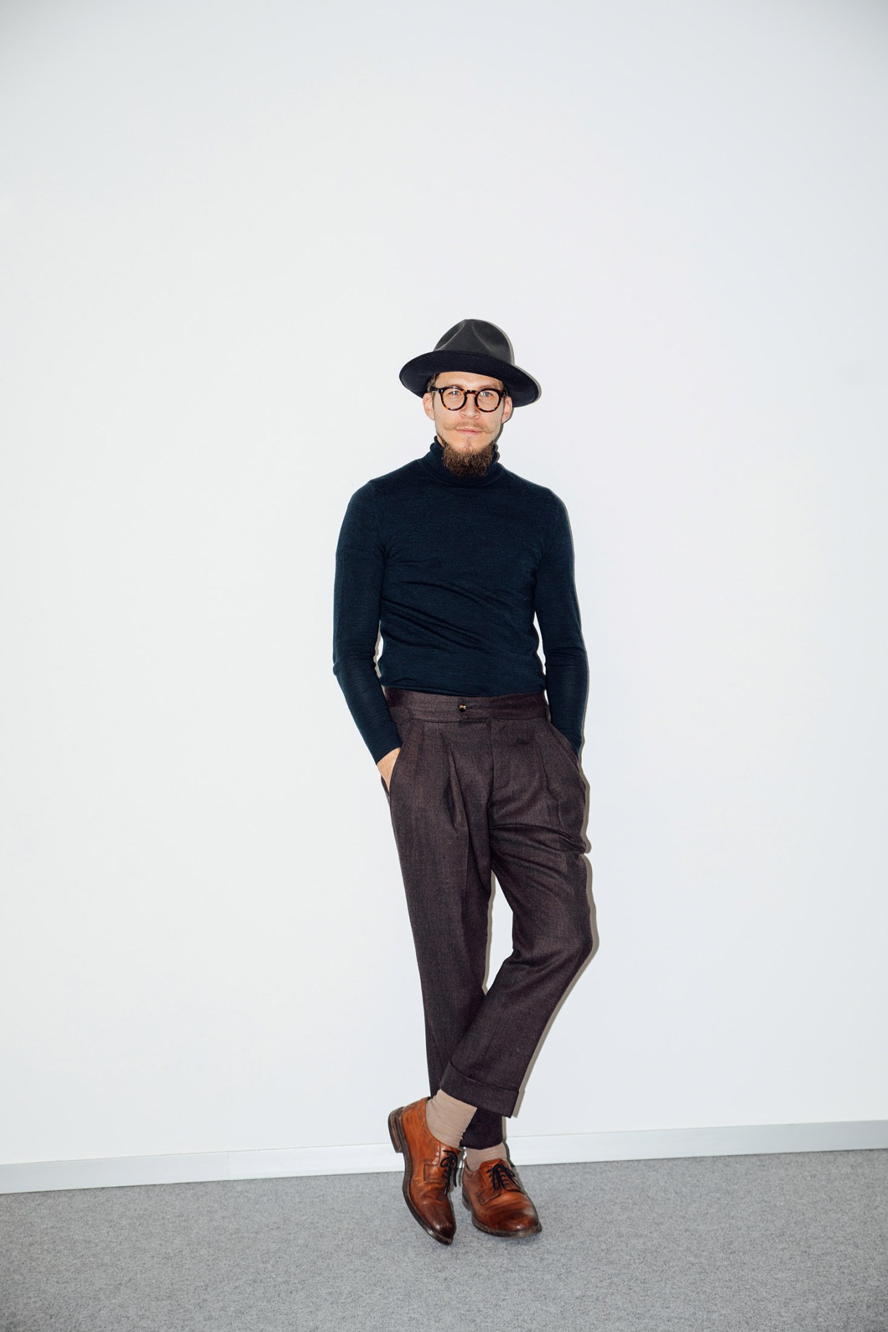 Man wearing dark turtleneck, charcoal trousers, black hat and brown shoes standing against white background.