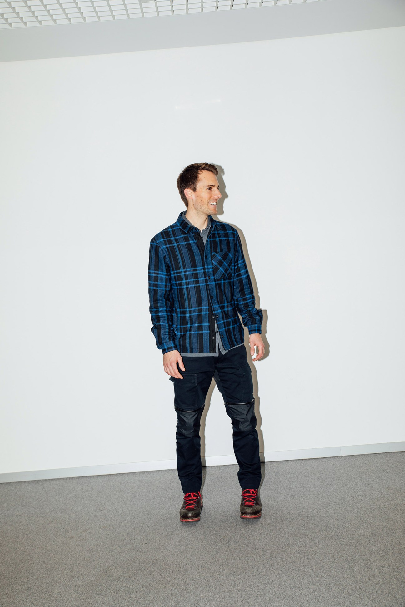 Man wearing blue plaid shirt, black pants with leather reinforcement panels, and red hiking boots stands against white wall.