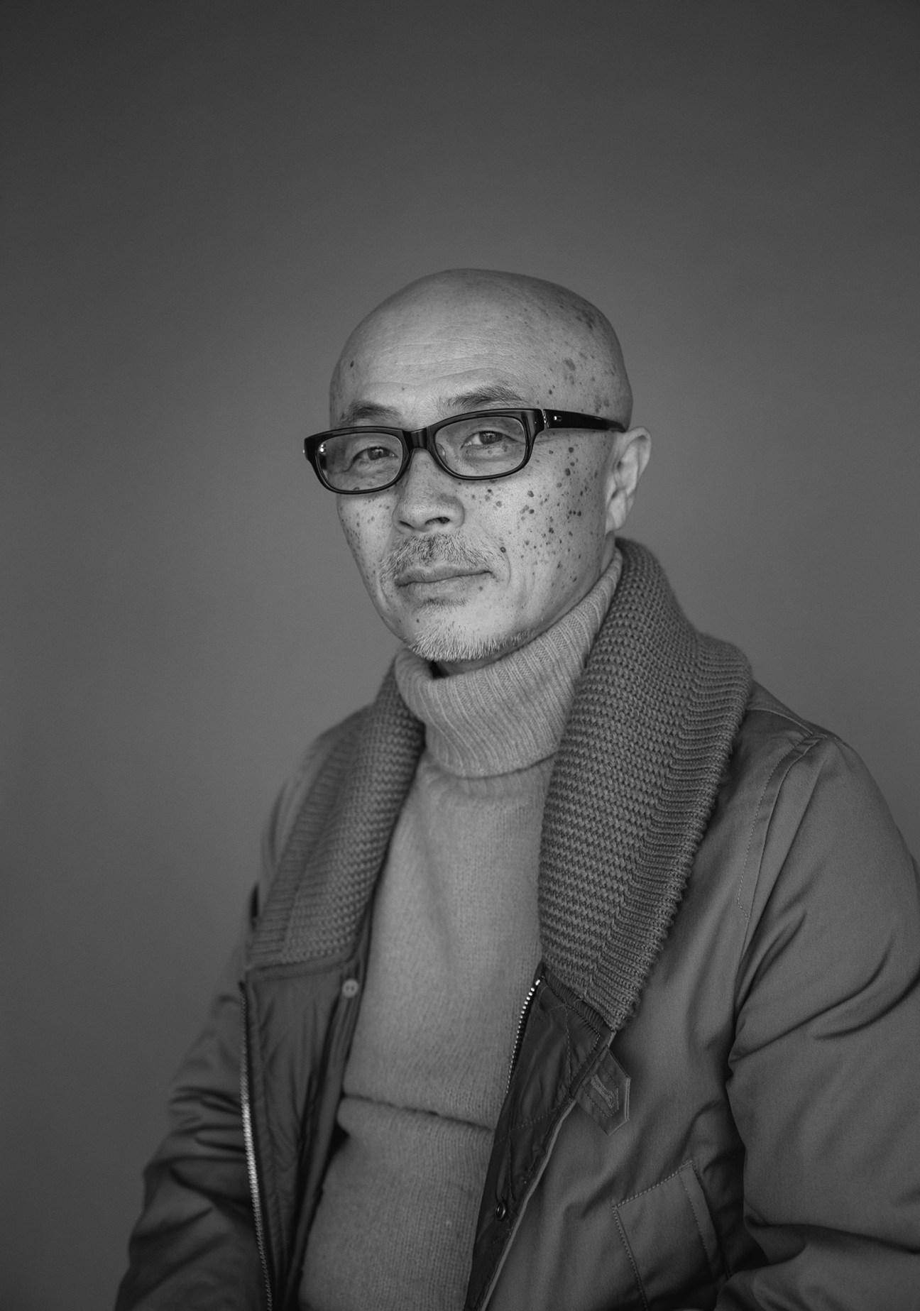 Black and white portrait of Eiichiro Homma, founder of Nanamica, wearing glasses and a layered jacket.